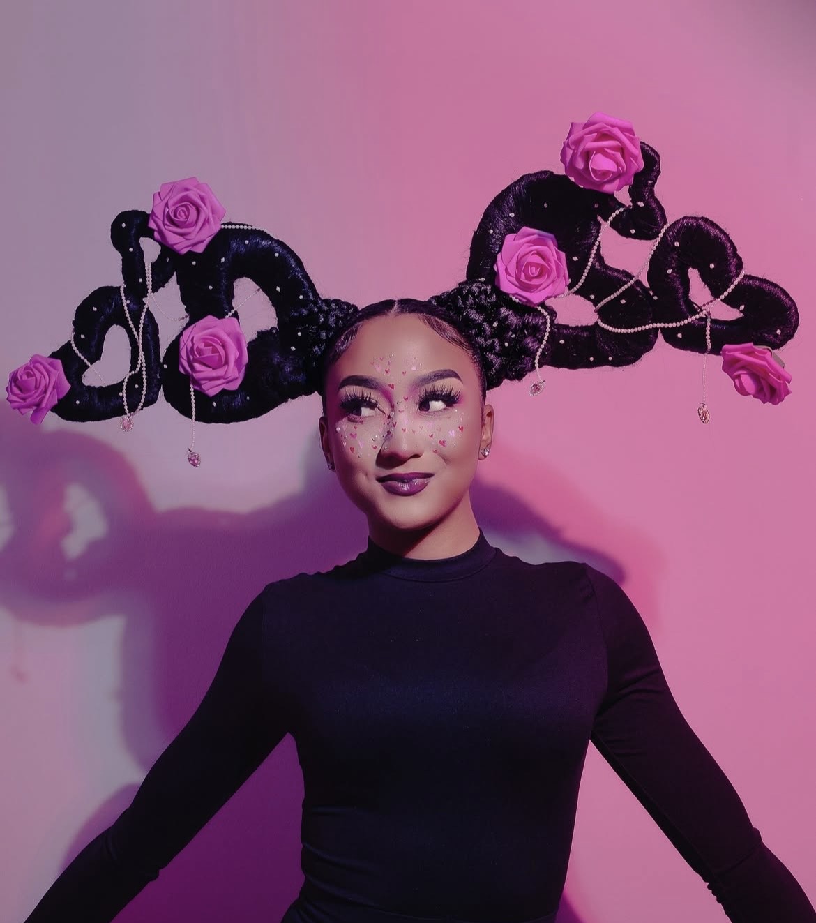 Young woman with styled hair and makeup, wearing a black top, standing in front of a pink background with artistic hair design and roses.