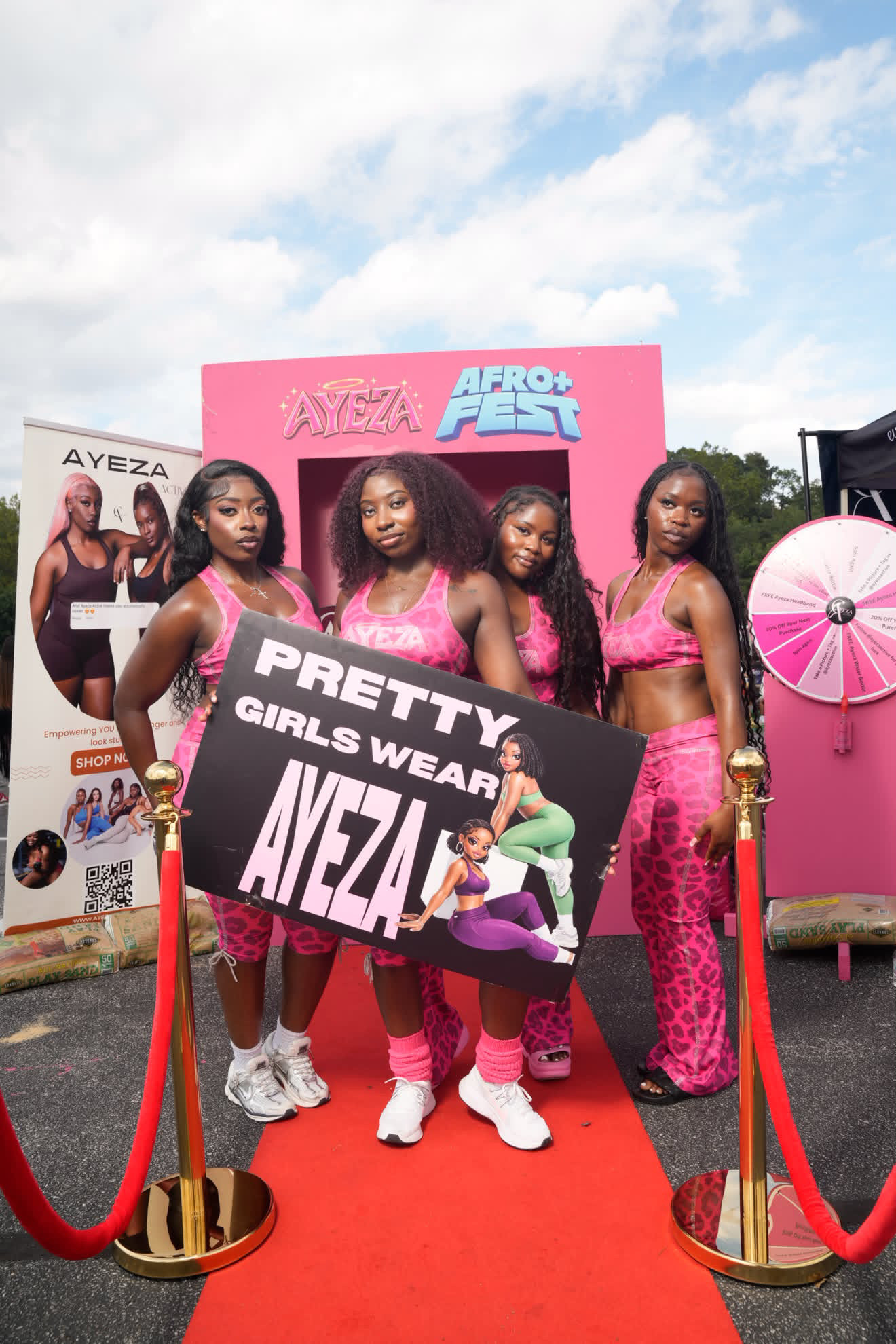 Four women in pink outfits holding a banner at an outdoor event with a pink backdrop and promotional posters.