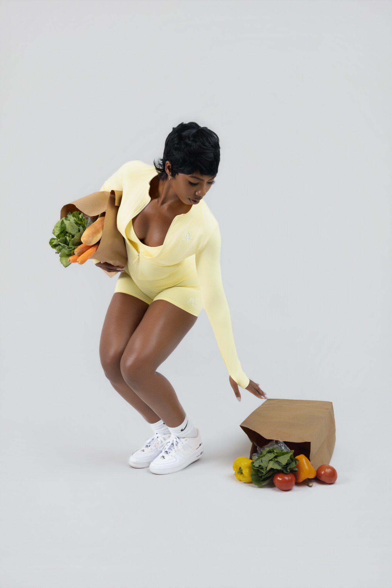 Person crouching, holding a bag of groceries with vegetables and fruits on the floor, against a plain background.