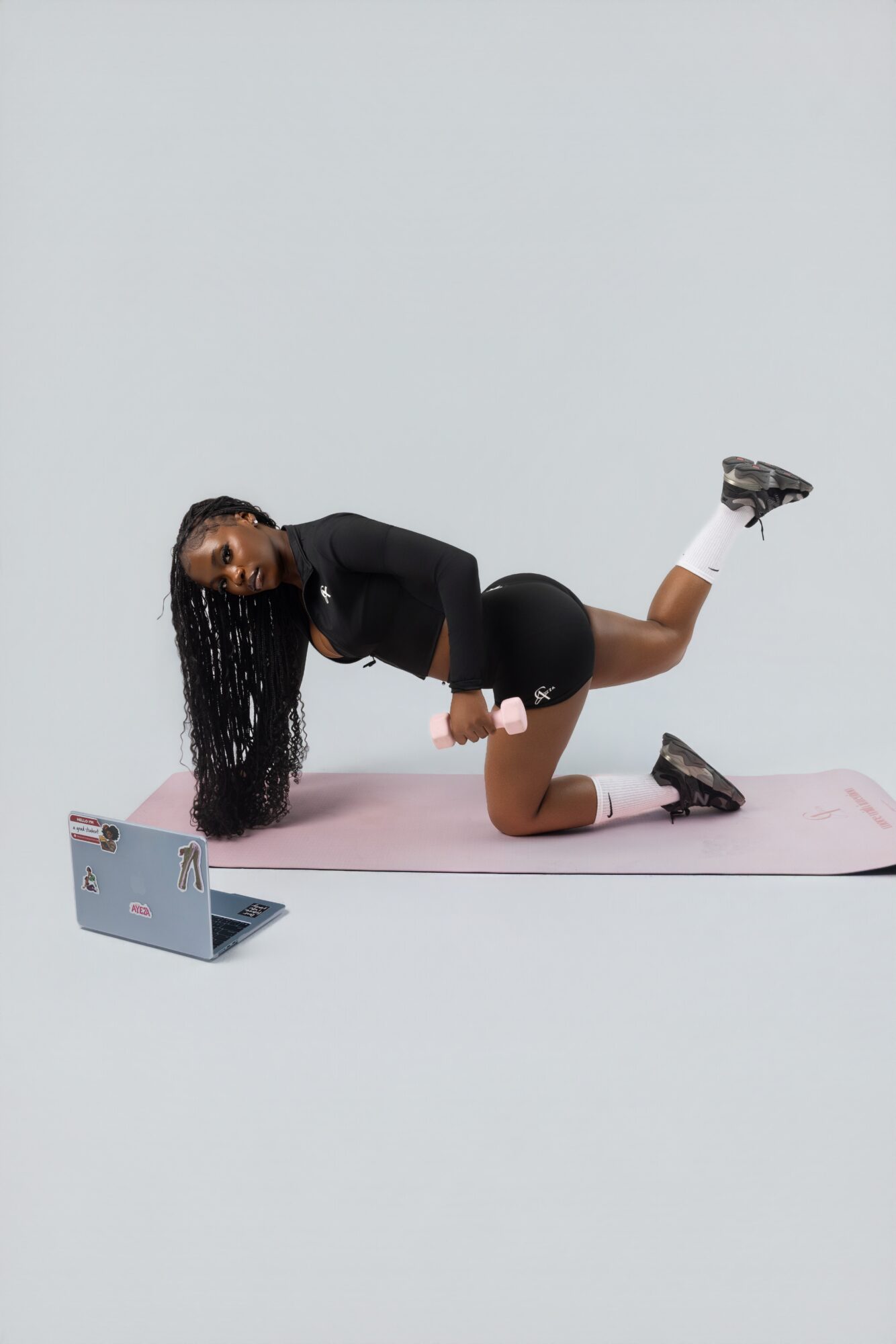Person in black sportswear on pink mat, supporting body with one knee and one hand, head turned to the side, against a plain background.