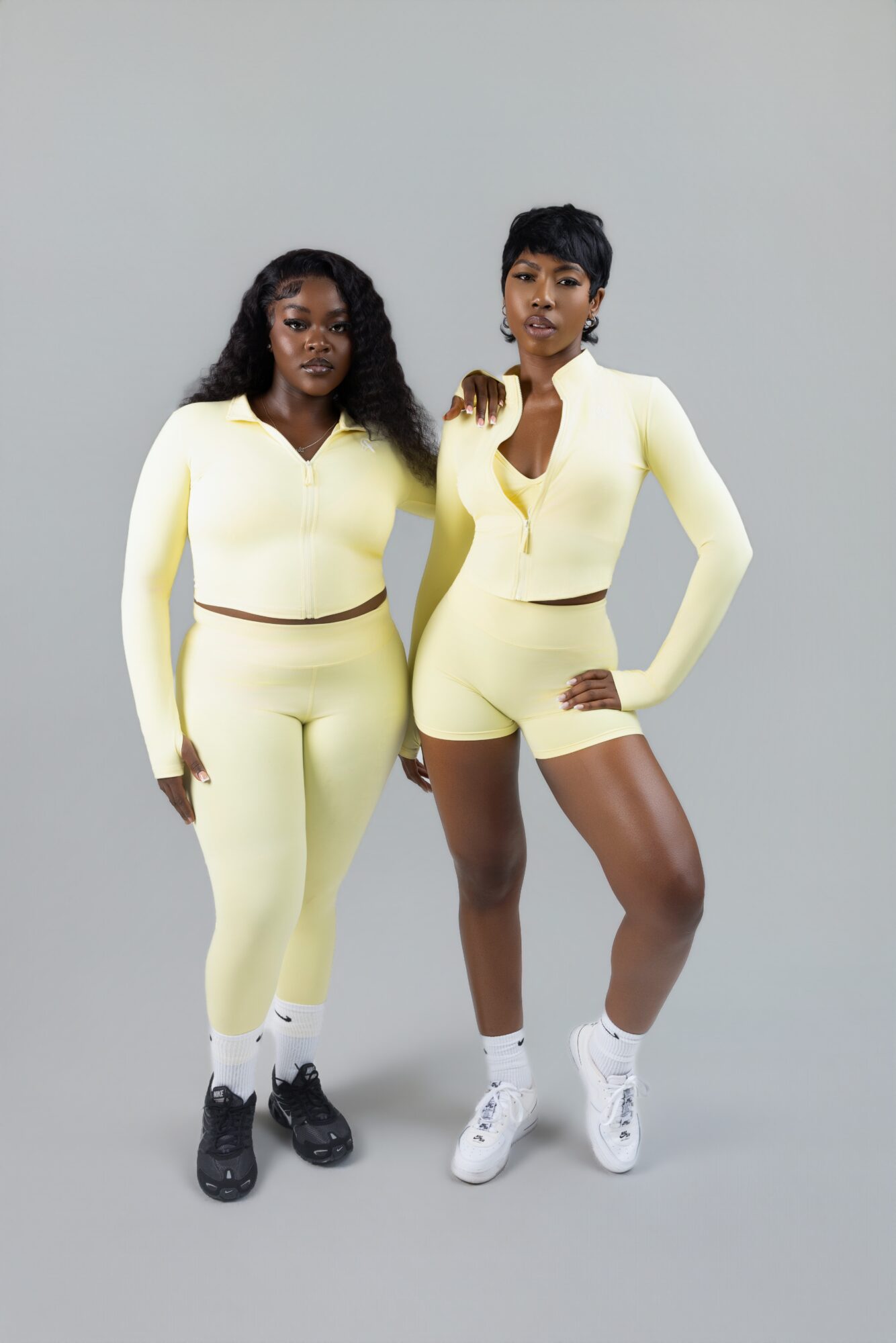 Two women stand side by side against a plain background, wearing matching yellow outfits and sneakers.