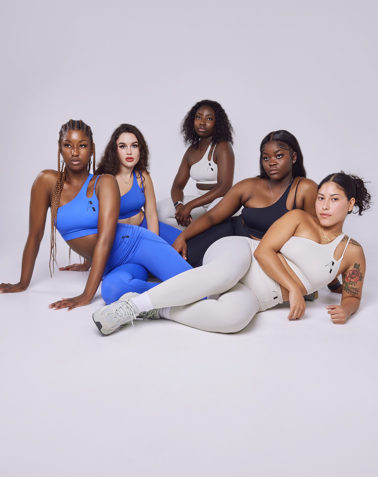 Five women pose together, some sitting and some lying down, wearing athletic clothing in white and blue, against a plain background.