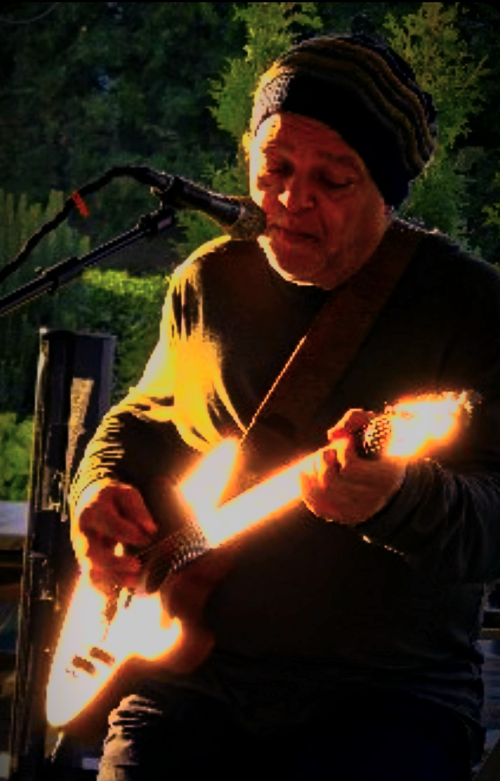 Person playing guitar outdoors with sunlight shining on the instrument, wearing a beanie and dark clothing.