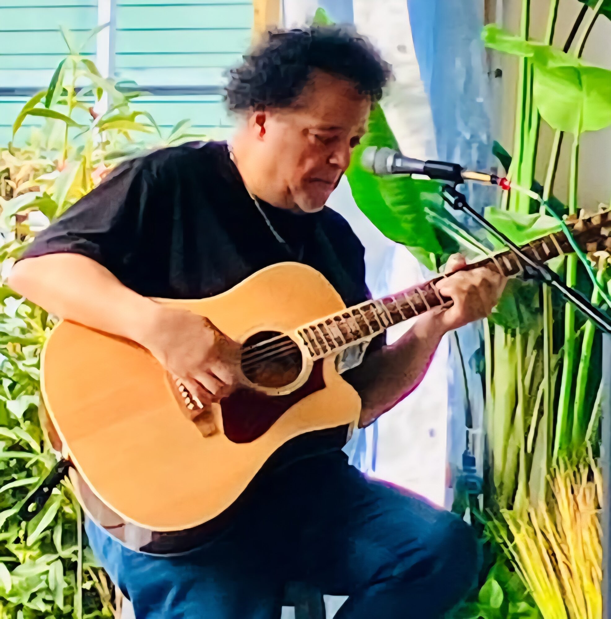 Person playing an acoustic guitar and singing into a microphone, surrounded by green plants, outdoors.