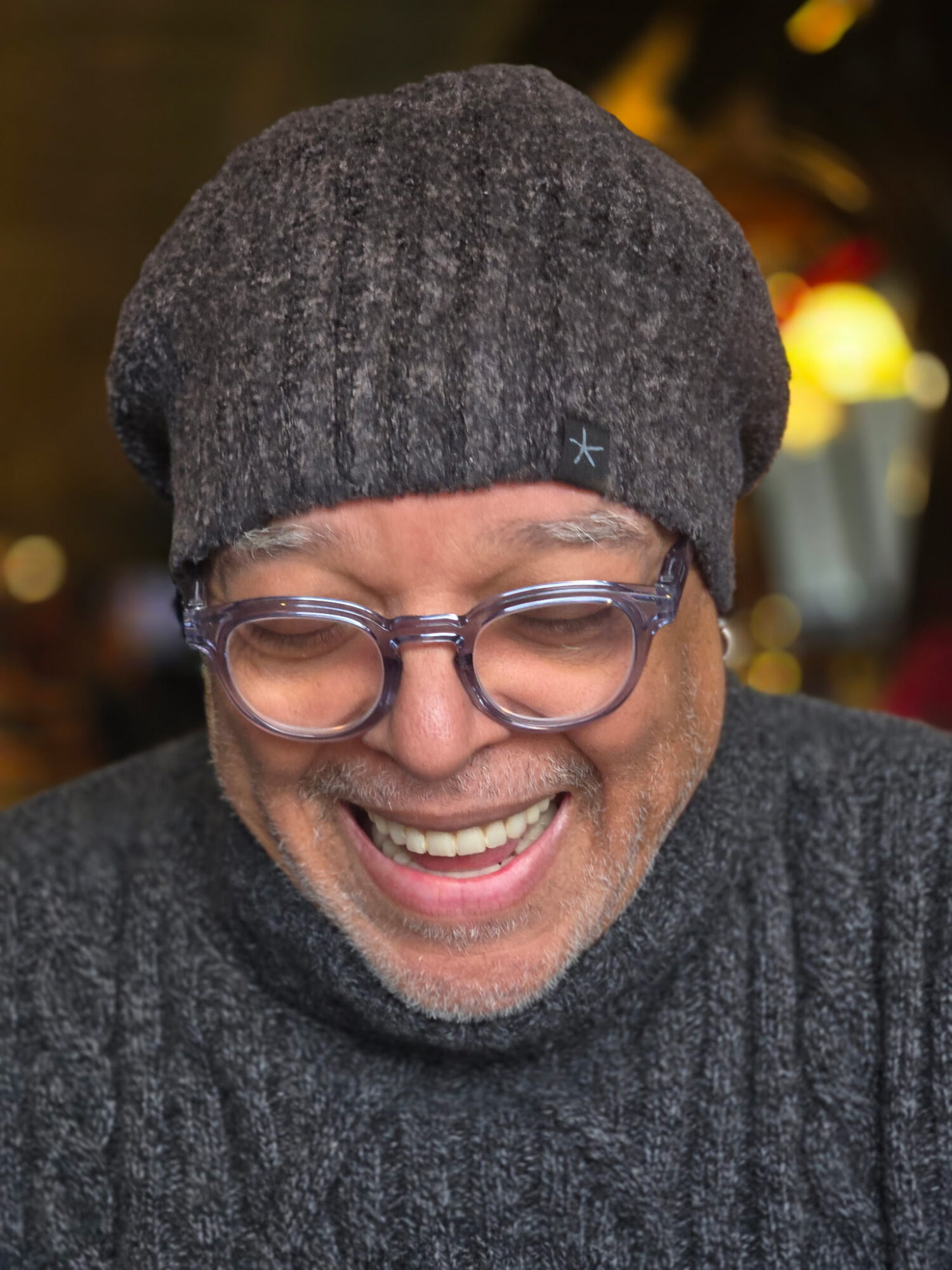 Man smiling, wearing glasses, a gray beanie, and a dark sweater, outdoors at night with blurred lights in background.