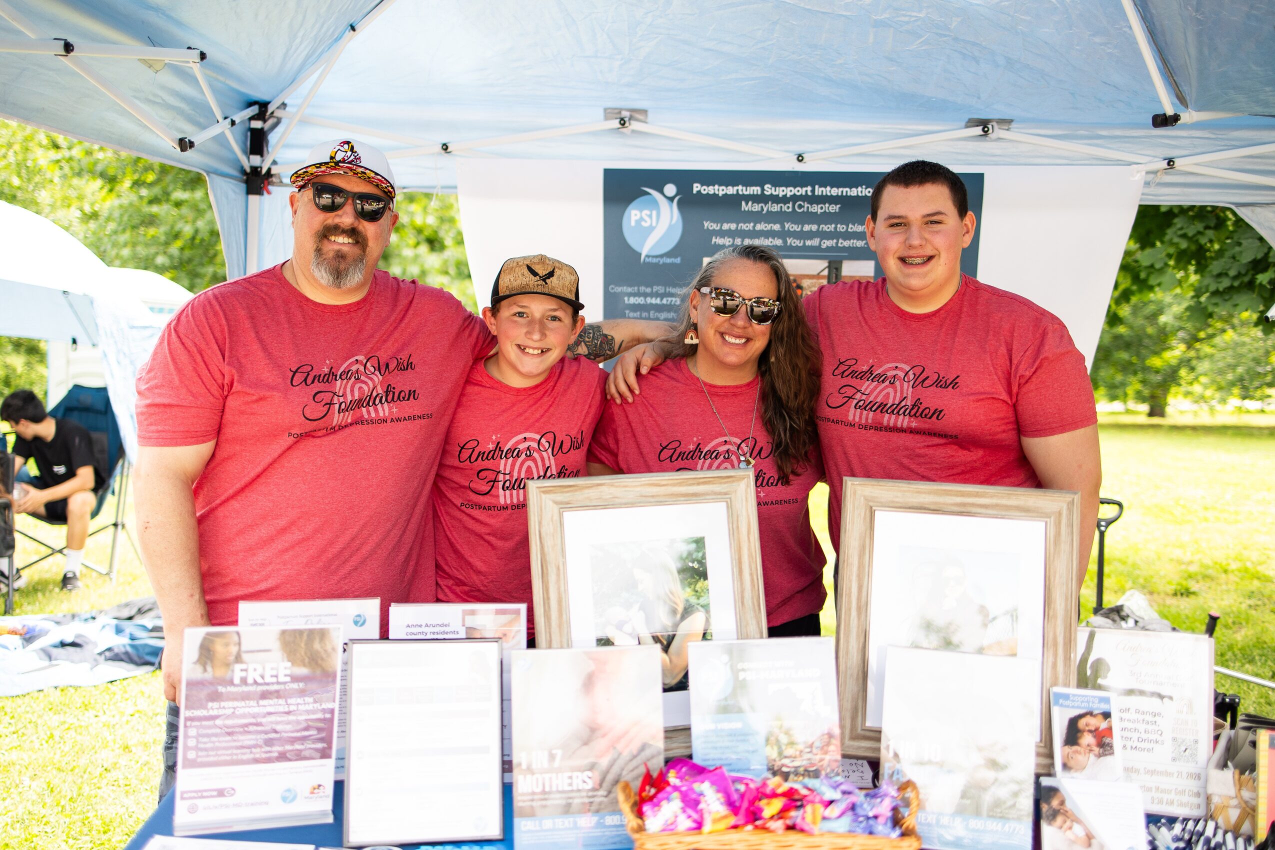 Four people wearing matching red shirts standing behind a table with framed photos and informational materials outdoors.