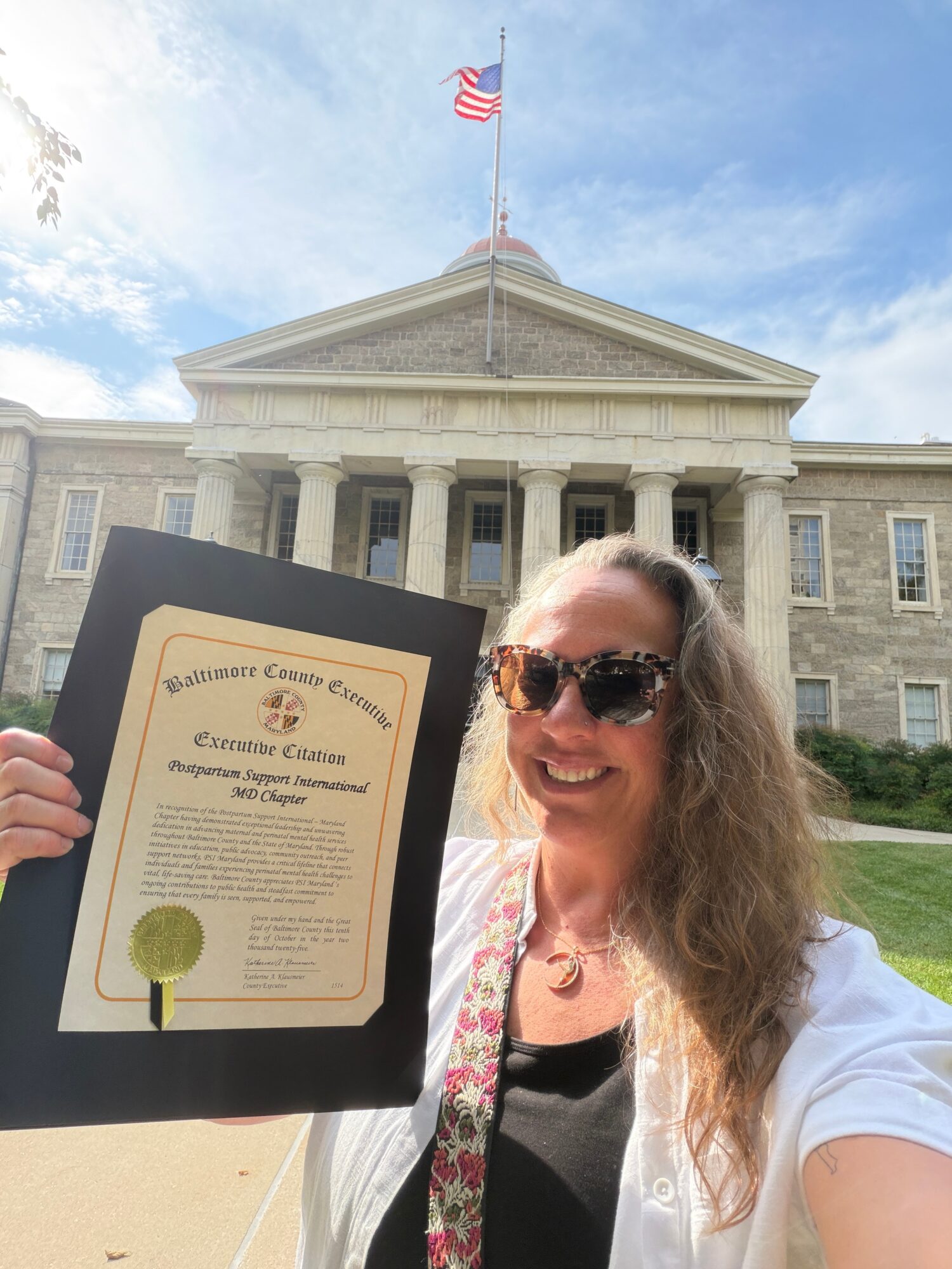 Woman with sunglasses holding a certificate in front of a large government building, blue sky, and American flag.