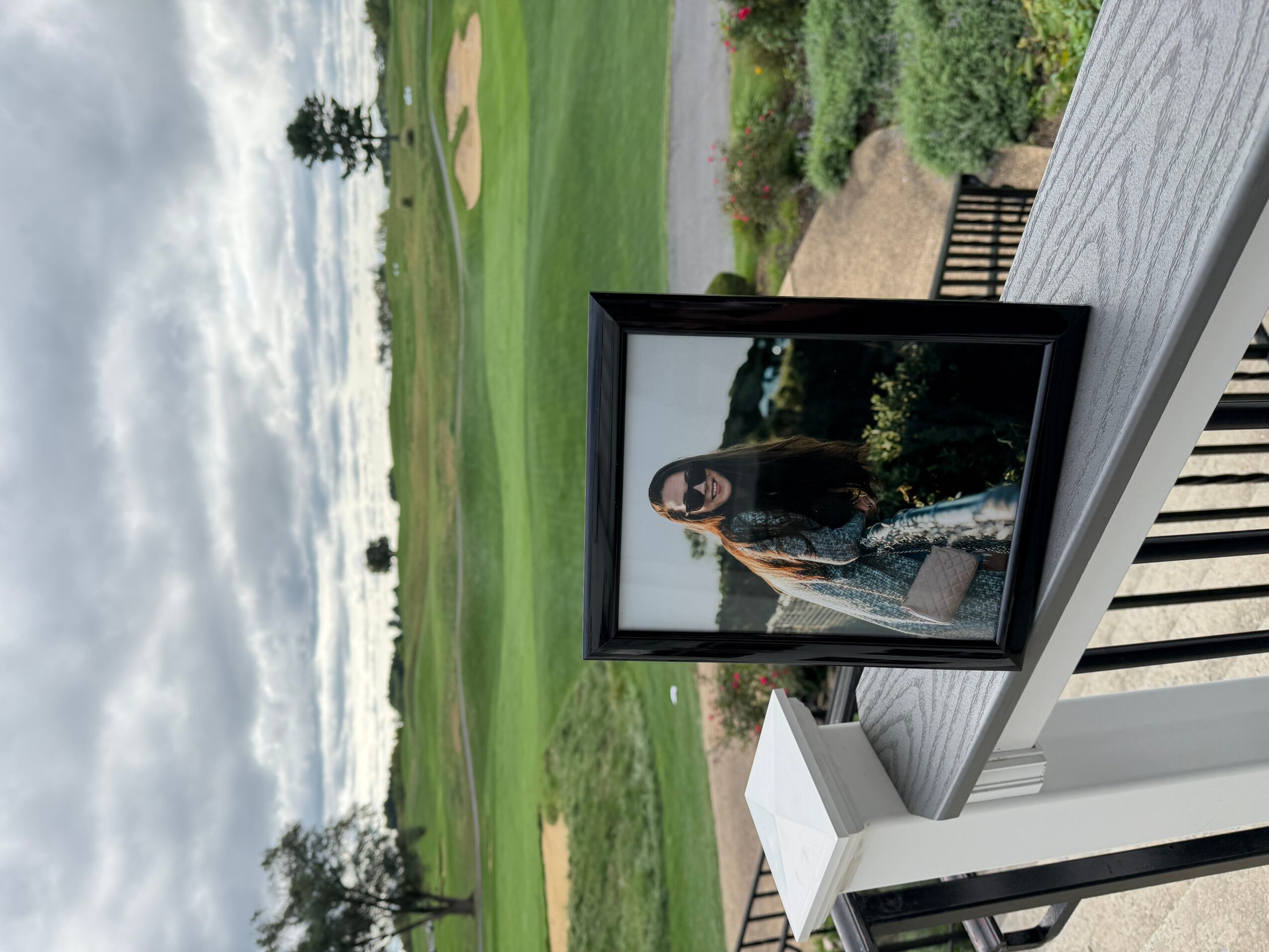Framed photograph of a woman with long dark hair, standing outdoors with trees and greenery in the background.
