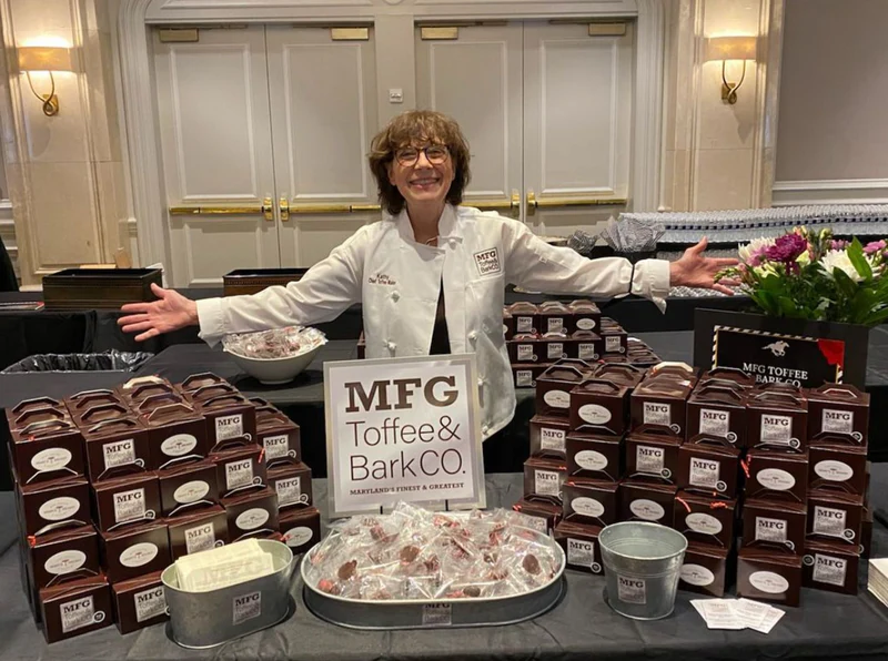 A person stands behind a table displaying stacked chocolate boxes, a platter of candies, and a sign for MFG Toffee & Bark Co.