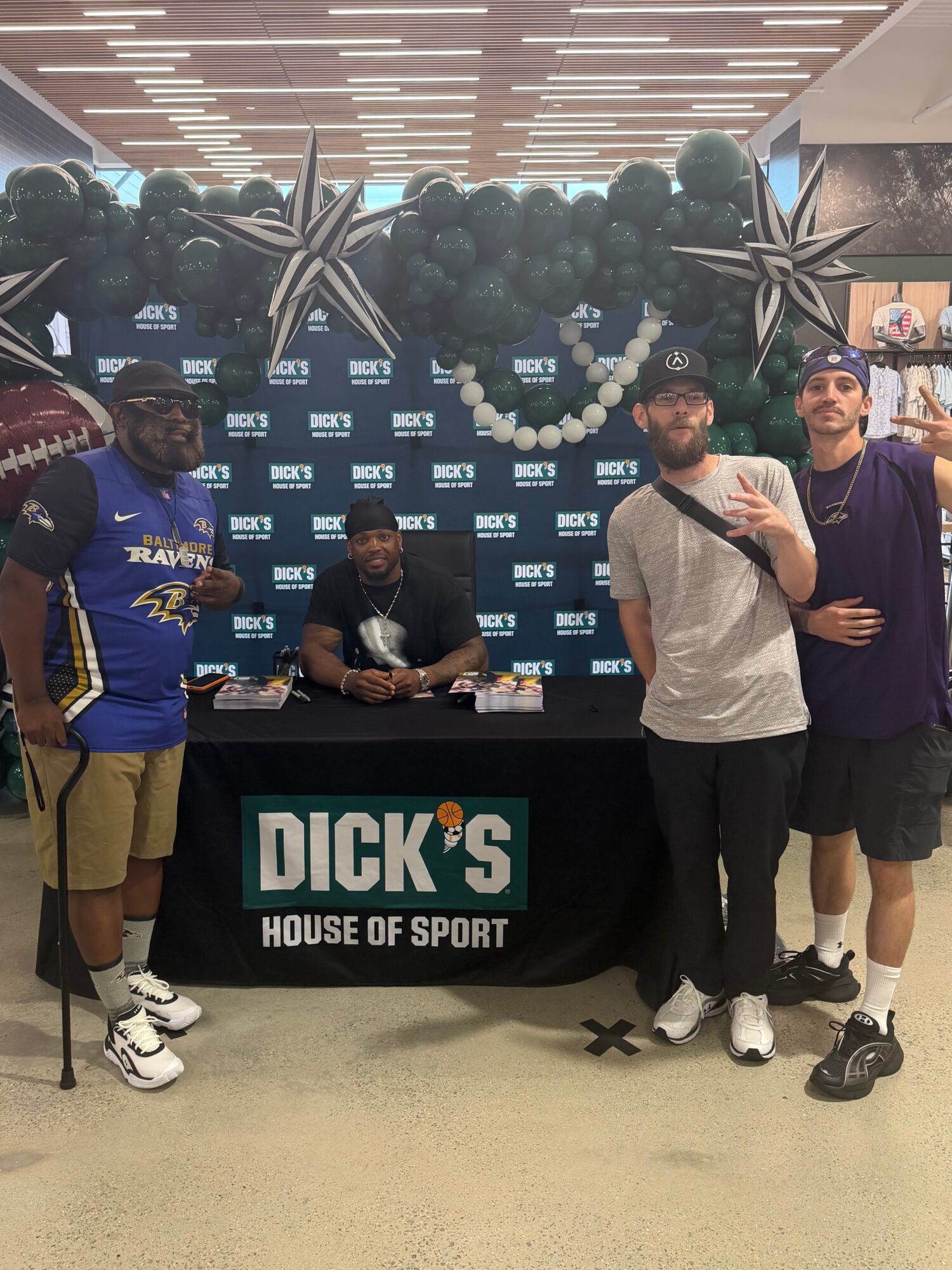Four men stand around a table with a Dick's House of Sport sign, balloons, and a backdrop, inside a store.