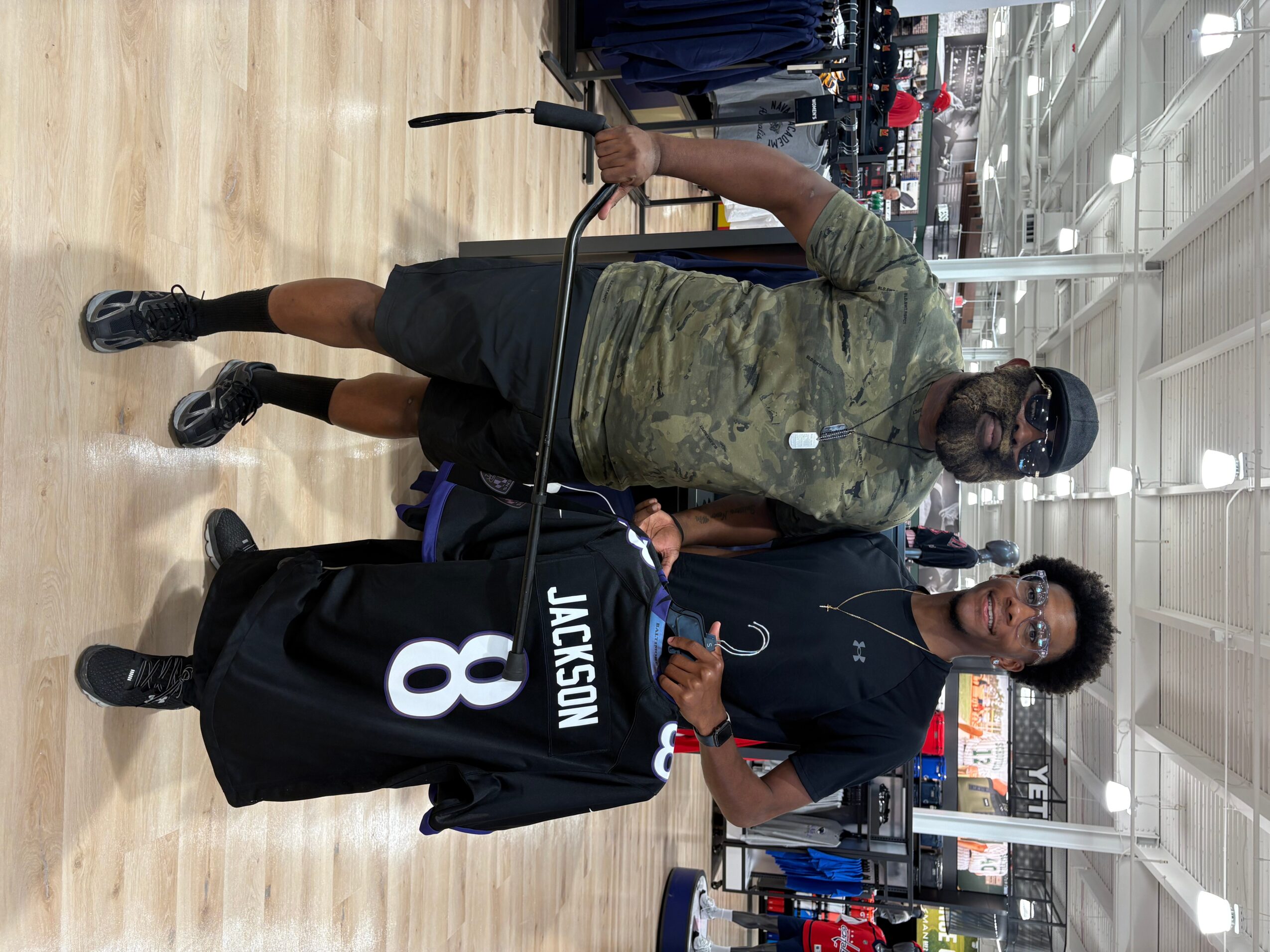 Two men standing in an indoor gym, one in camouflage shirt and shorts, the other in black sportswear, with gym equipment around.