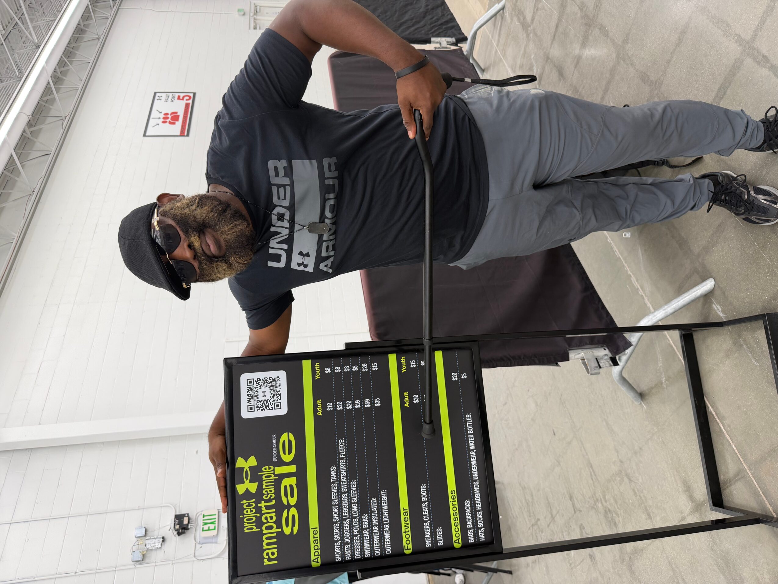 Person standing next to a promotional sign in an indoor setting, wearing a black cap and gray pants, holding a black bag.