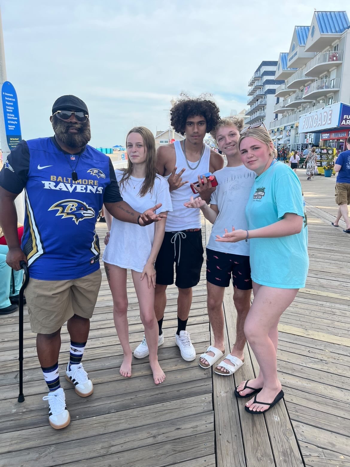Group of five people standing on a boardwalk near buildings and shops, some holding drinks, with a cloudy sky above.