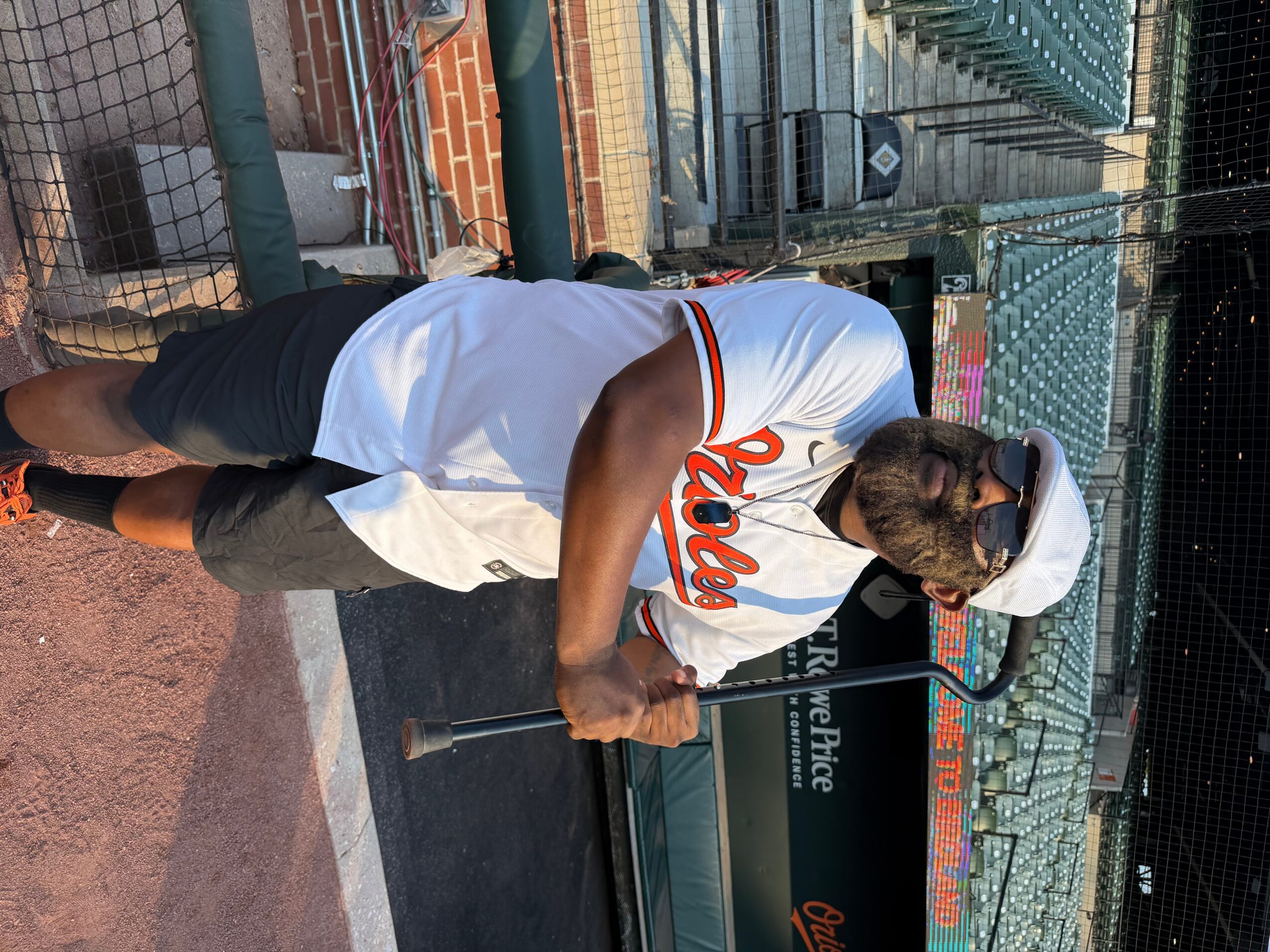 Person wearing a white cap and white shirt with orange and black text, holding a baseball bat on a baseball field.