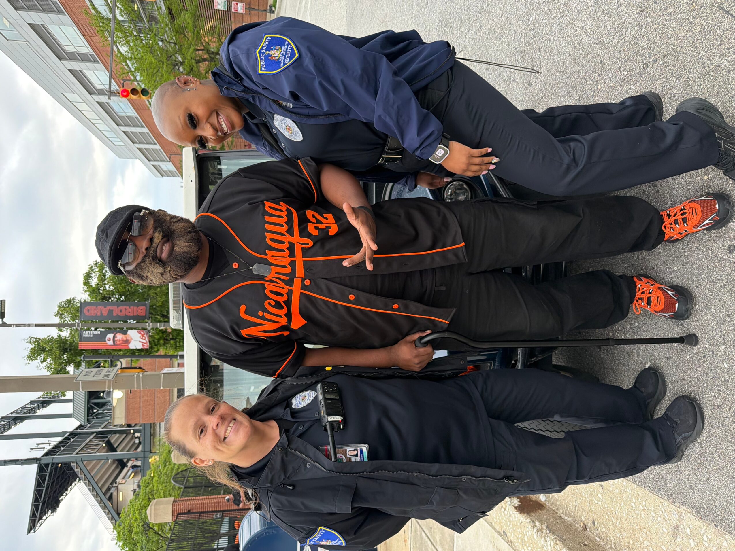 Three people standing outdoors on a sidewalk, two in police uniforms and one in casual clothing, holding a sign.