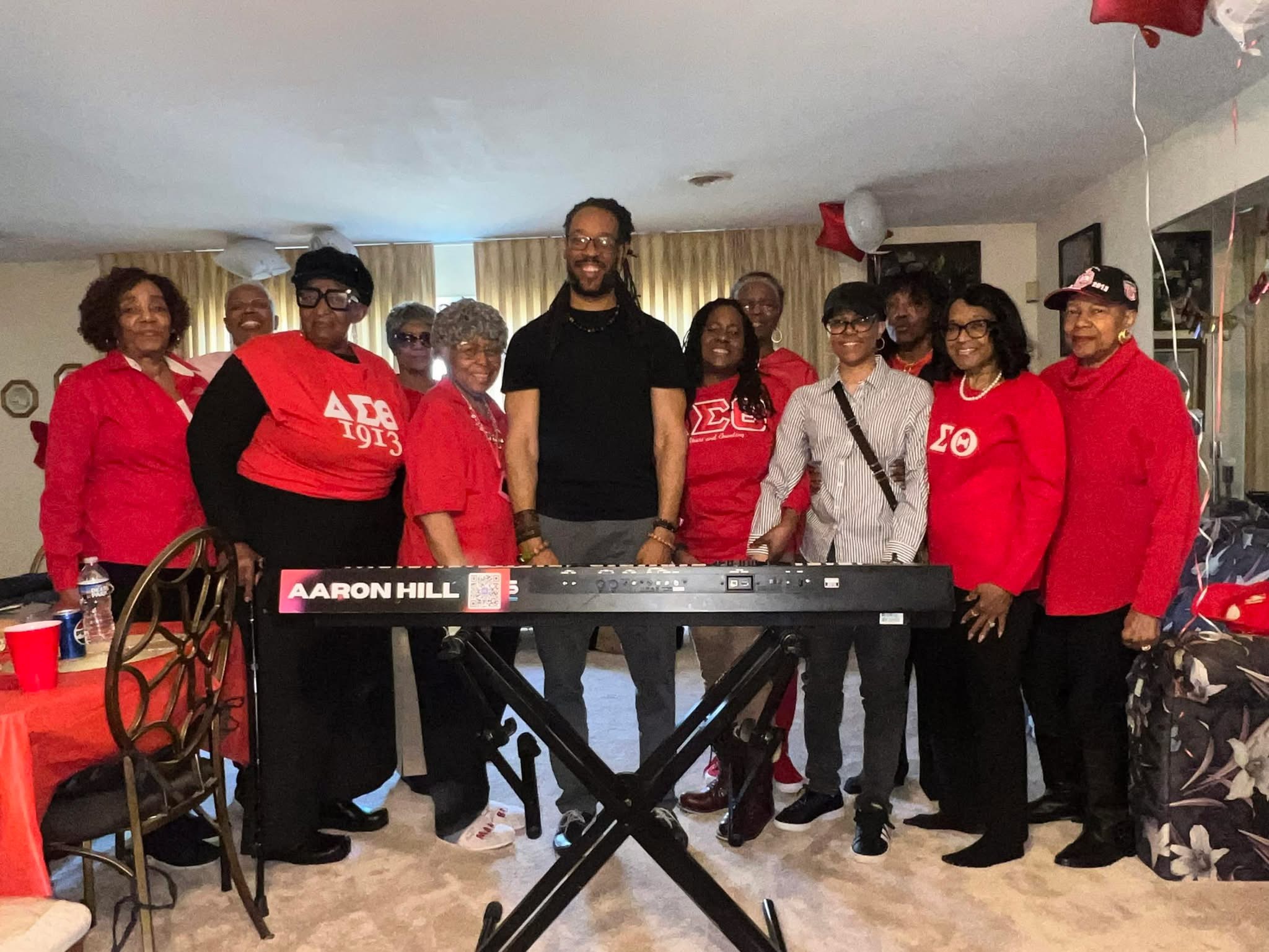Group of ten people standing indoors, some wearing red clothing, with a keyboard in front, celebrating together.