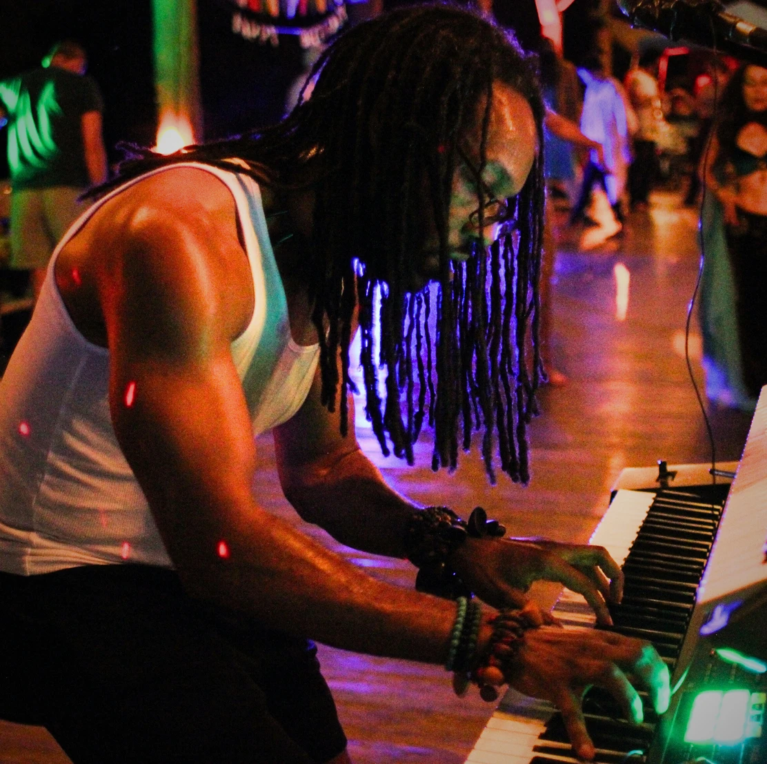 Person with long dreadlocks playing keyboard in a dimly lit, colorful environment.
