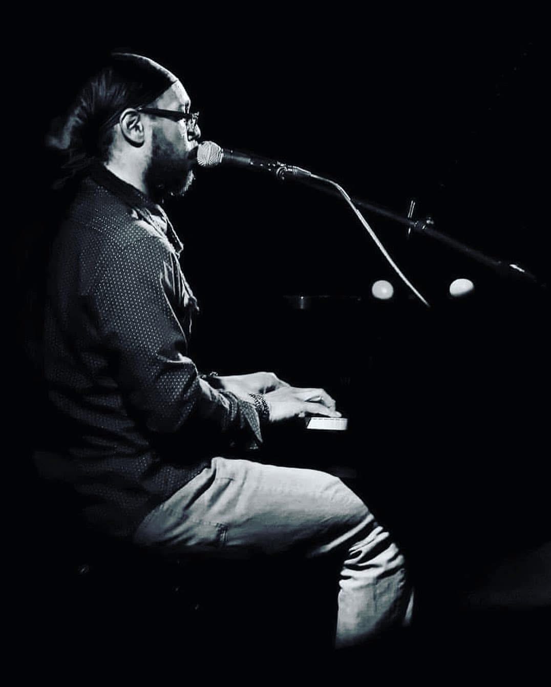 Person playing keyboard and singing into microphone on stage, dark background, black and white image.