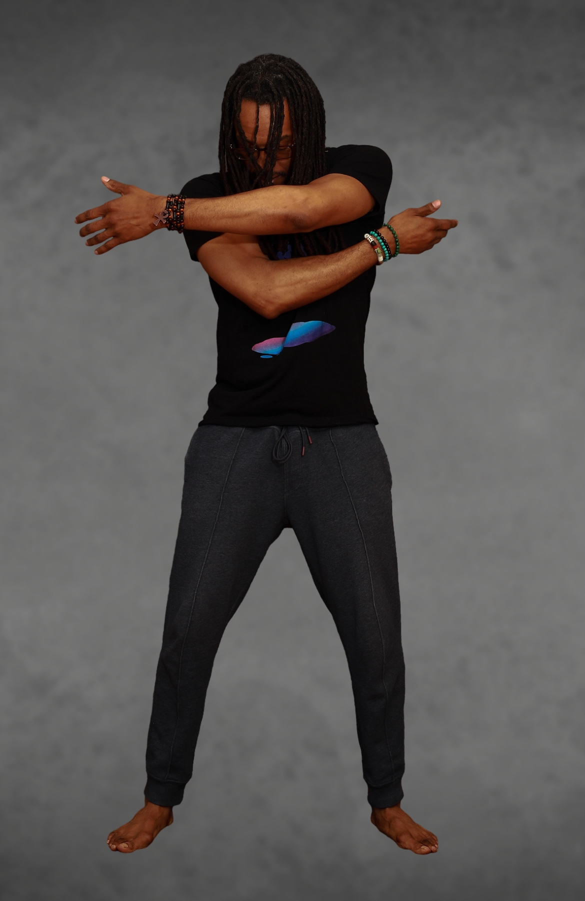 Person standing with arms crossed in front, wearing a black shirt and dark pants, against a gray background.
