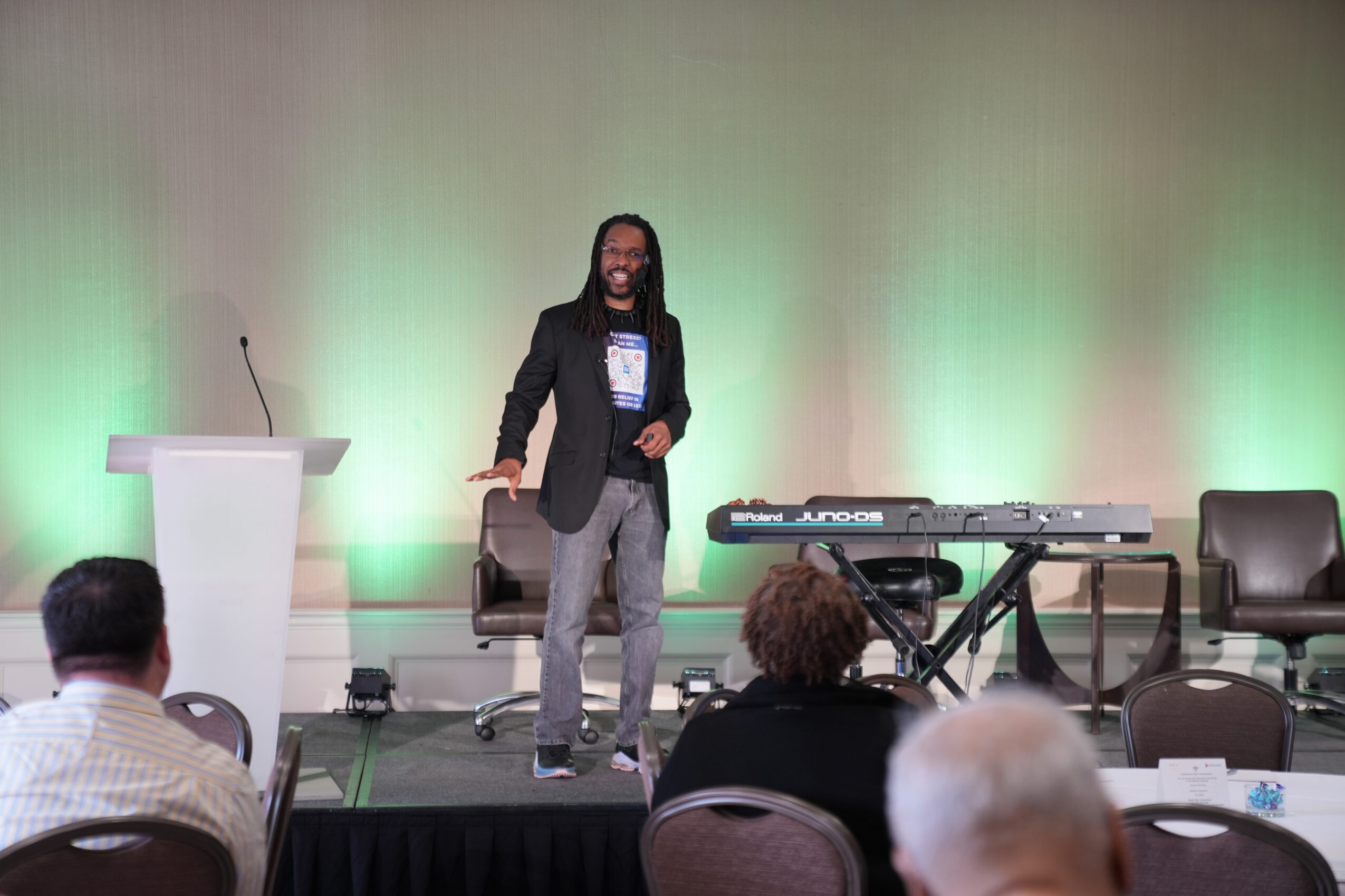 A man stands on stage with a microphone, a keyboard, and chairs, speaking to an audience in a conference room.