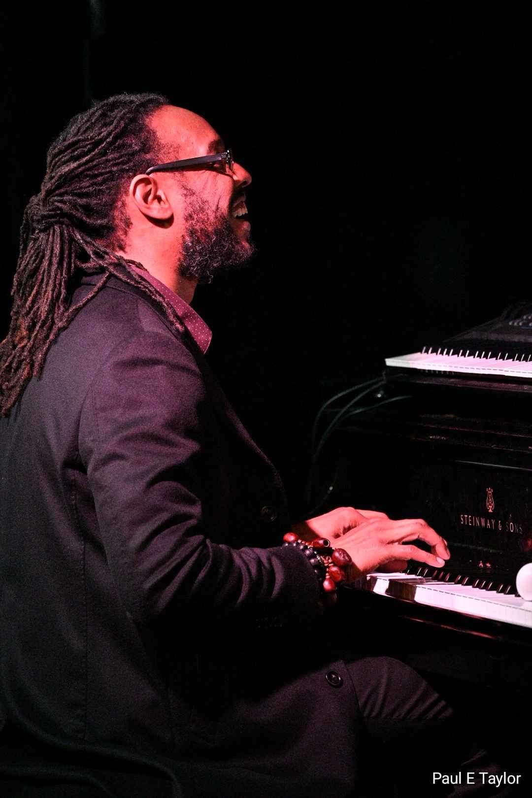 Man with dreadlocks and glasses playing a piano on stage, dark background, side view, smiling, wearing dark clothing.