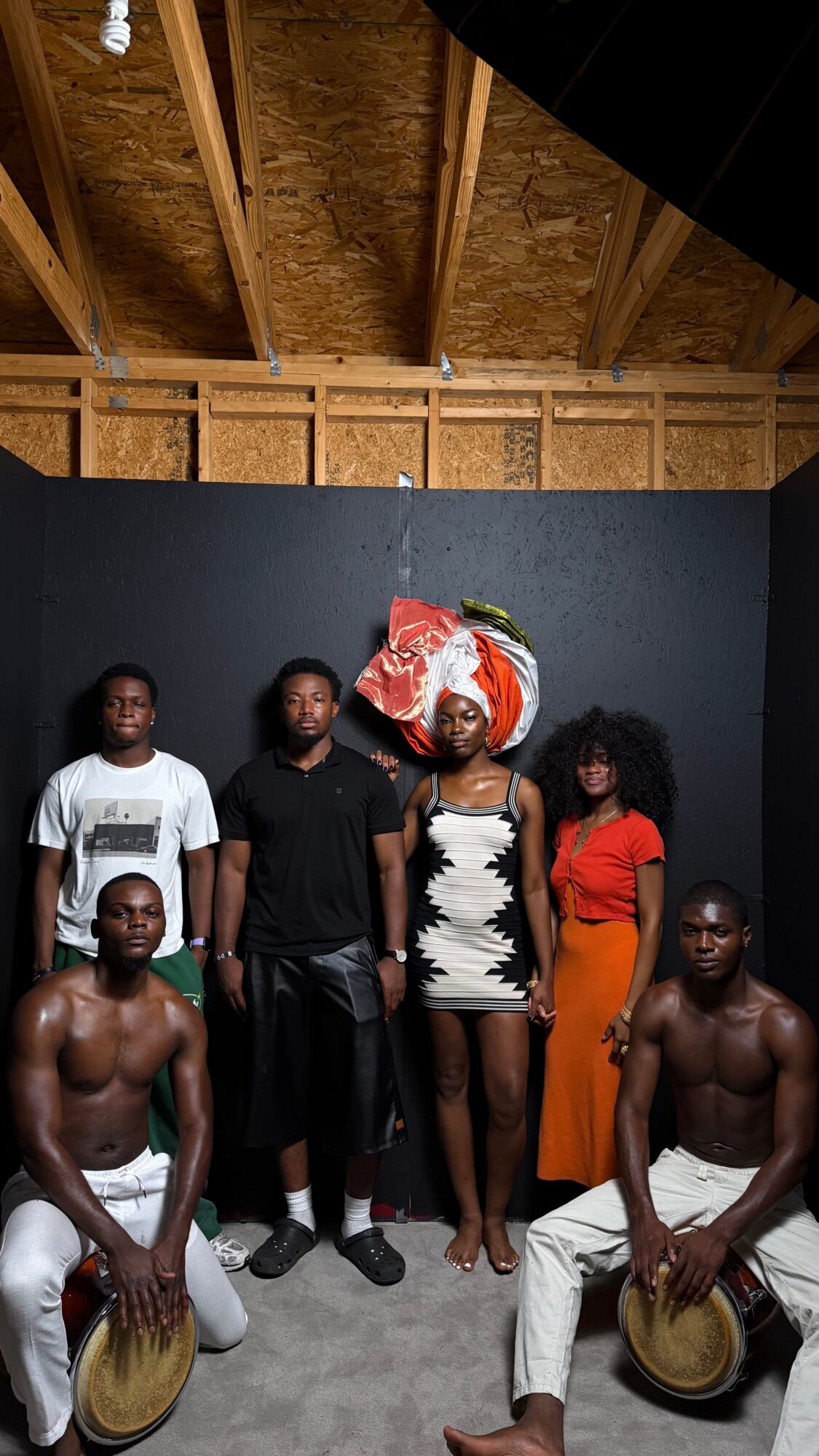 Group of six people, some holding drums, standing in front of a black wall with a wooden ceiling above.