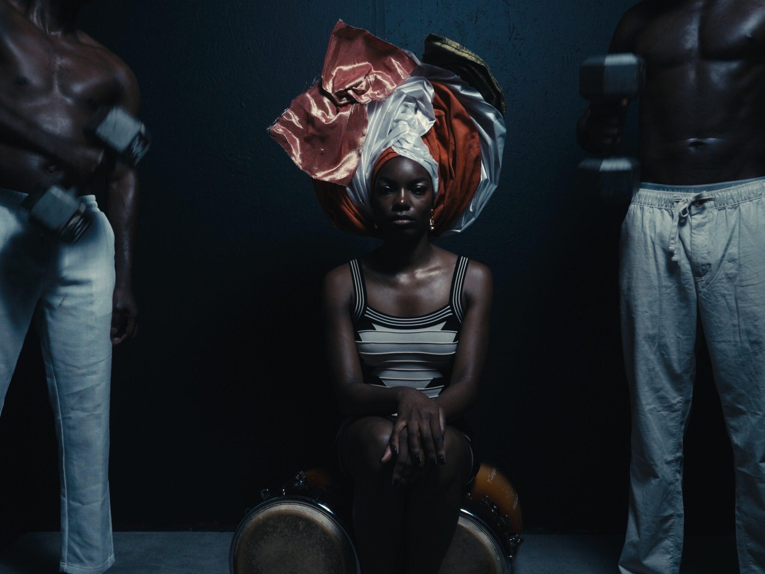 Young girl with elaborate headwrap sitting with hands clasped, surrounded by shirtless men against dark background.
