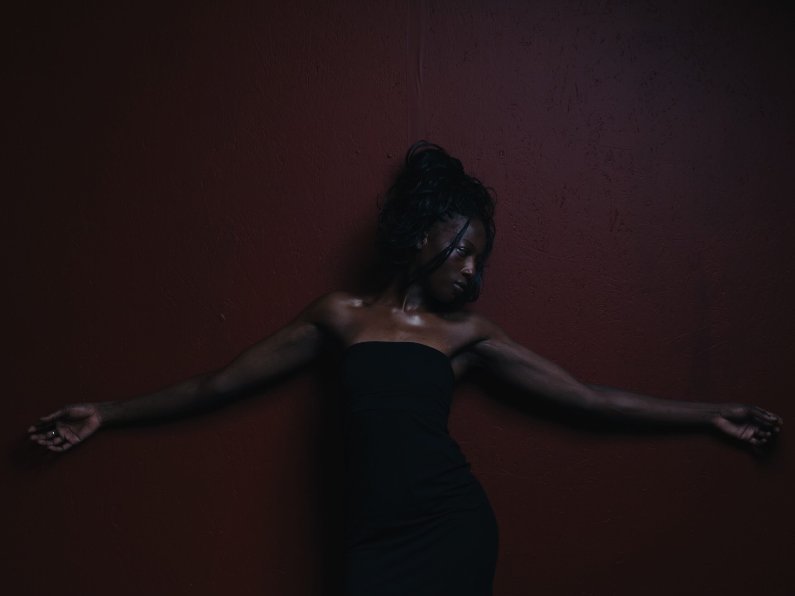 Person in a black dress leaning against a dark red wall with arms outstretched, head tilted downward, hair in a bun.