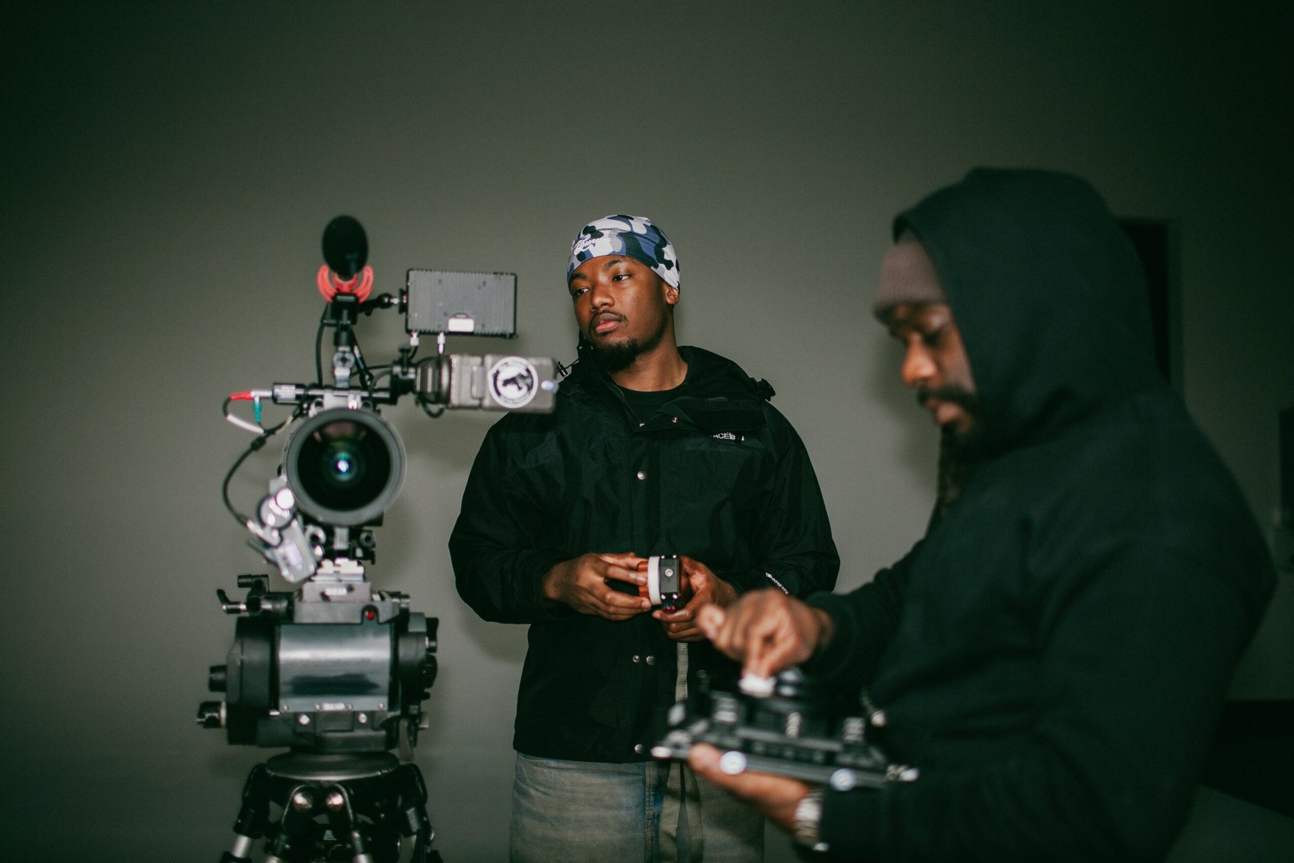 Two men in dark clothing, one with a headband, stand near a professional camera setup in a dimly lit room.