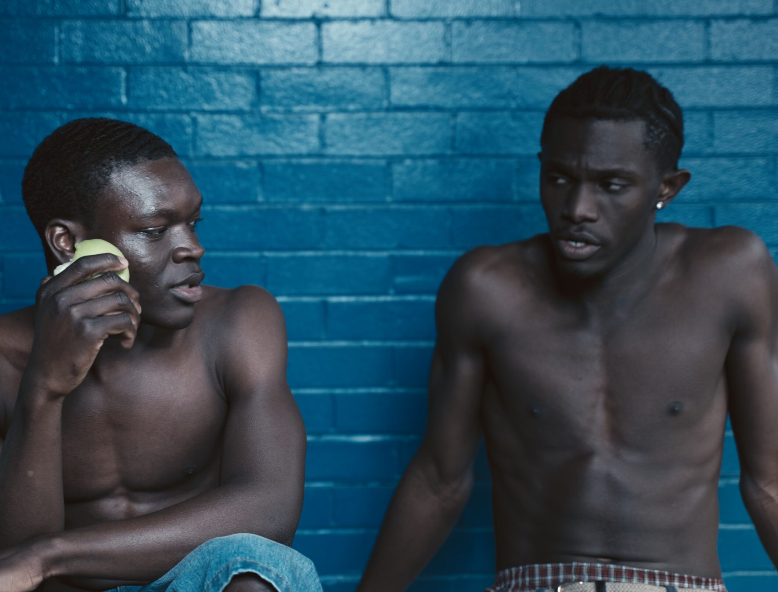 Two shirtless men sit against a blue brick wall, one holding a phone to his ear, the other looking at him.