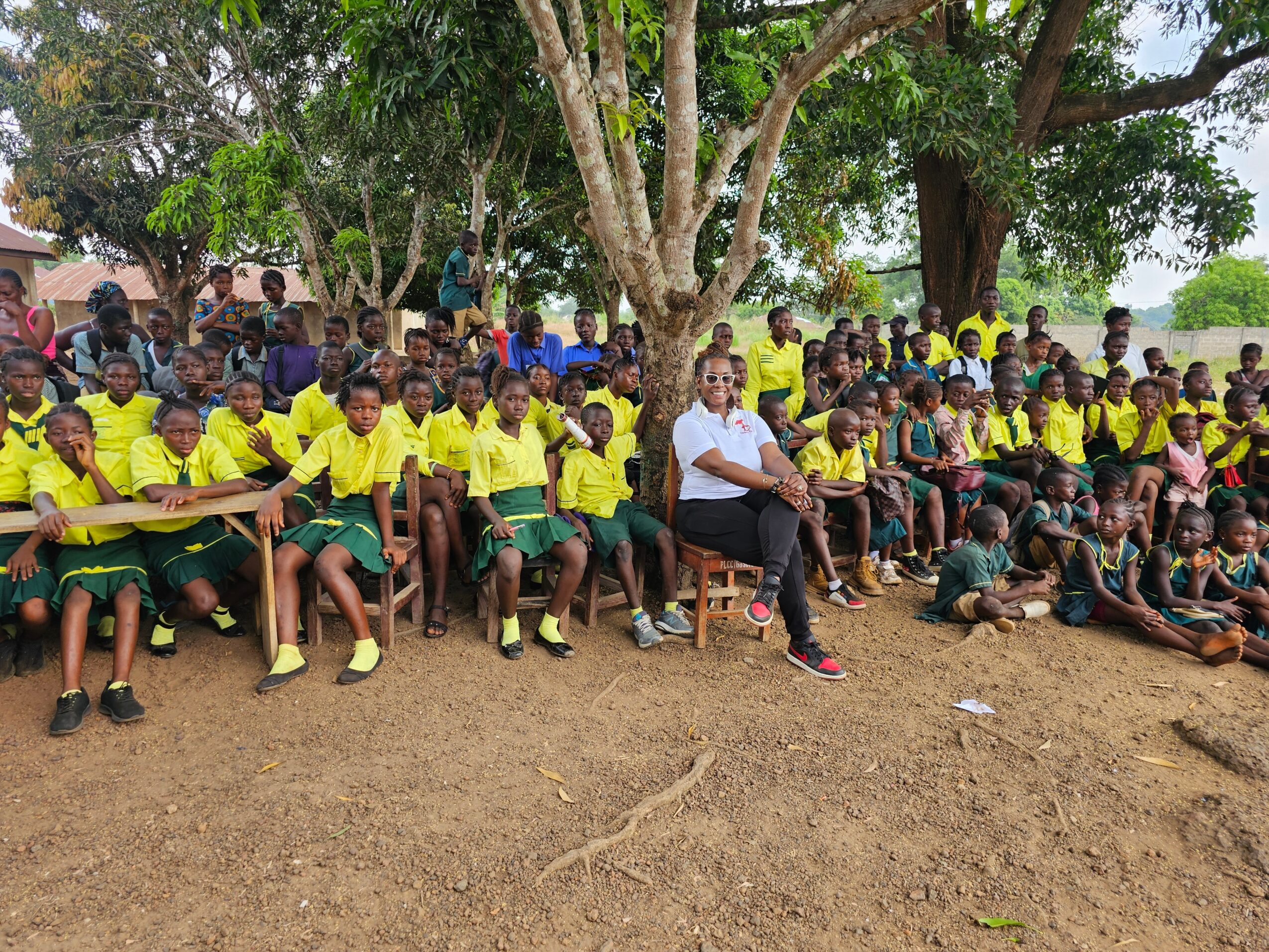 Group of children and a woman sitting outdoors under a large tree, some children wearing yellow shirts, others in darker clothing.