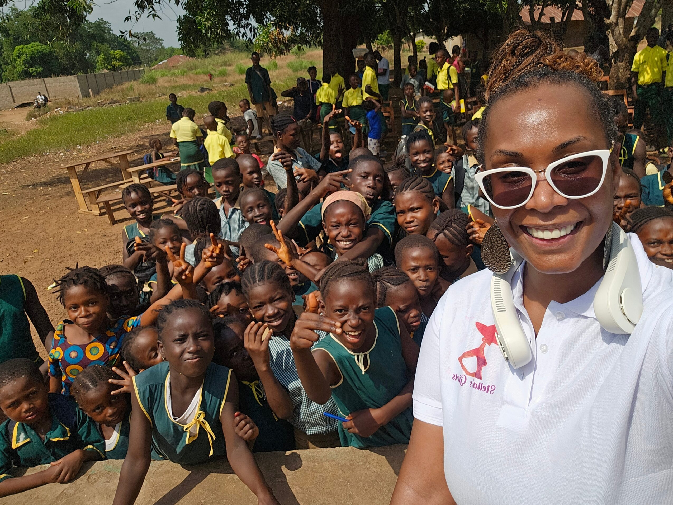 A woman with sunglasses and headphones takes a selfie with a large group of children outdoors, some smiling and waving.