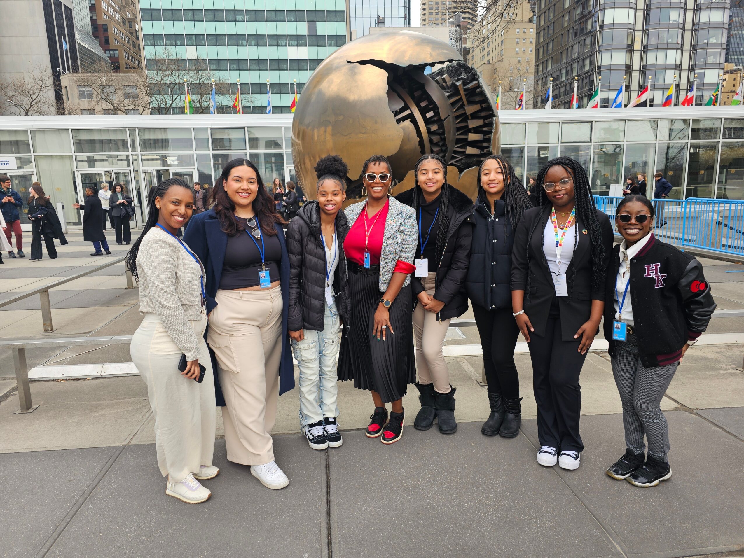Group of seven women standing outdoors in front of a large metallic sculpture in an urban area, smiling at the camera.