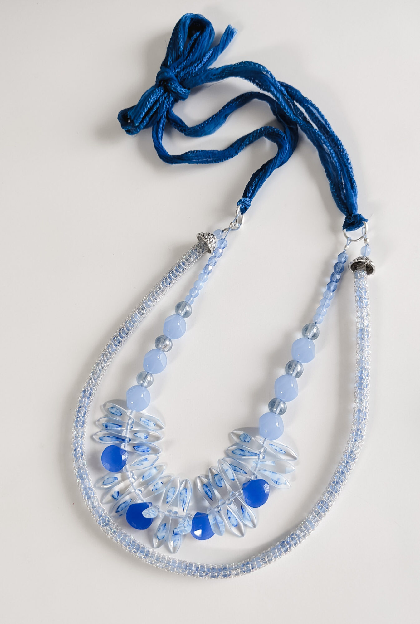 Blue fabric tie with beaded necklace featuring clear and blue beads, arranged in a semi-circle, on a white background.