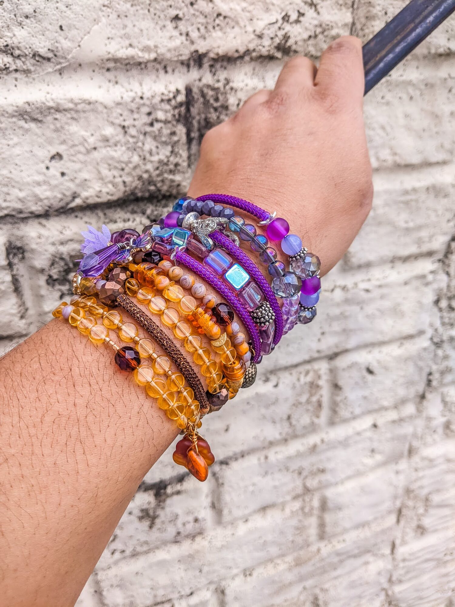 Hand with multiple colorful beaded bracelets against a brick wall background.