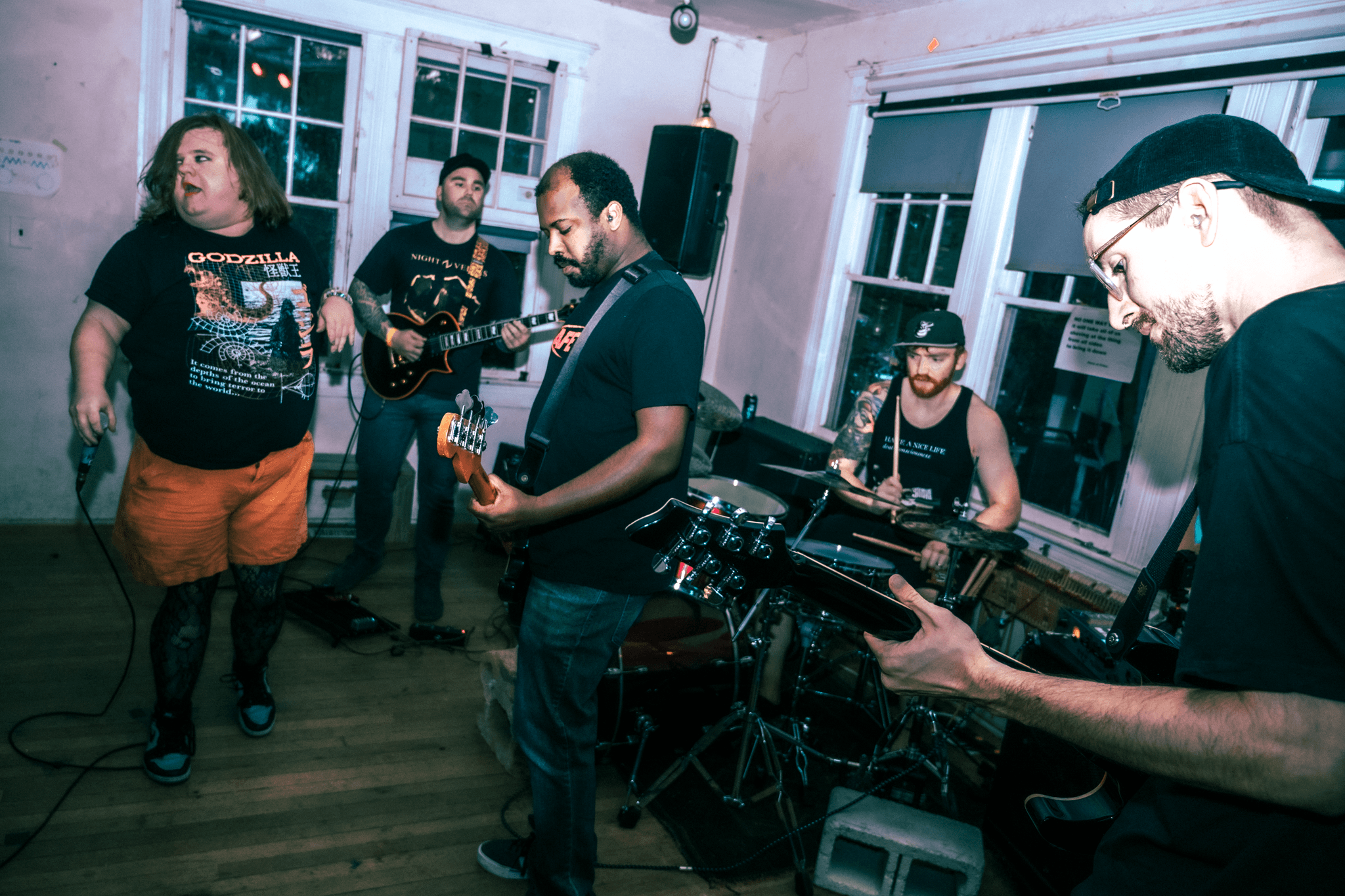 Group of five musicians performing indoors, with a woman singing and four men playing instruments, in a room with large windows.