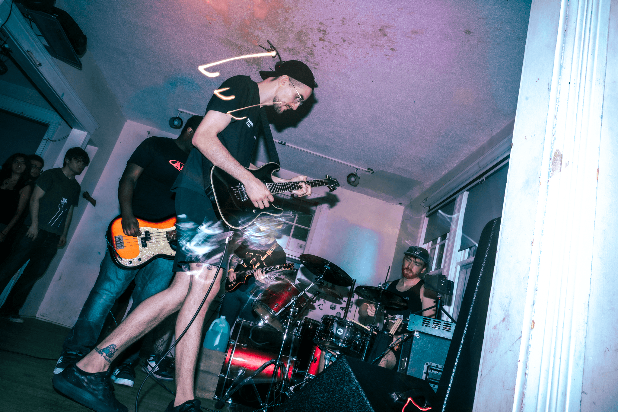 Musicians perform live with one playing guitar and another on drums in a room with audience members watching.
