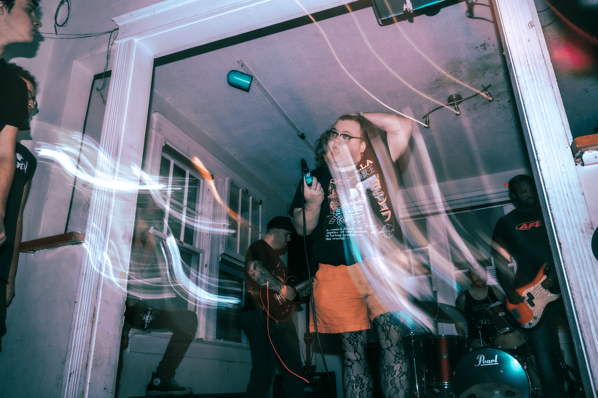 A woman singing into a microphone with a guitar and bass player in a room with glass walls, colorful light streaks.