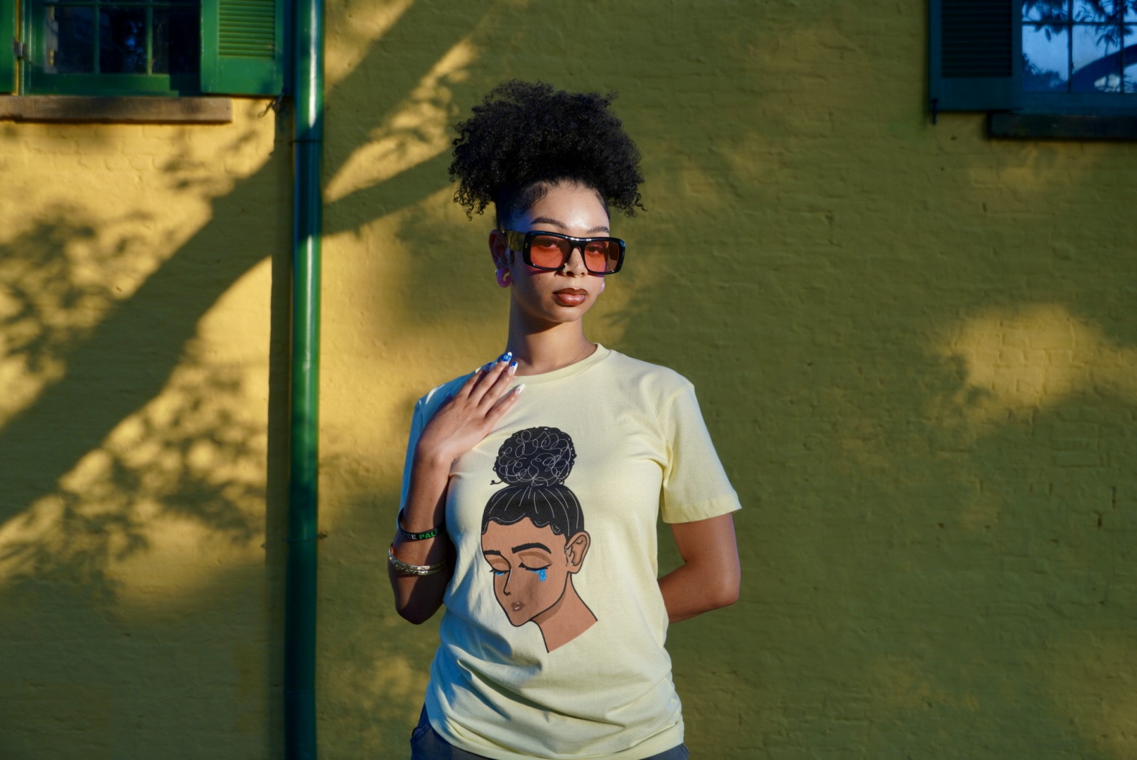 Young woman with sunglasses and curly hair stands against a yellow wall with shadows, wearing a graphic t-shirt.