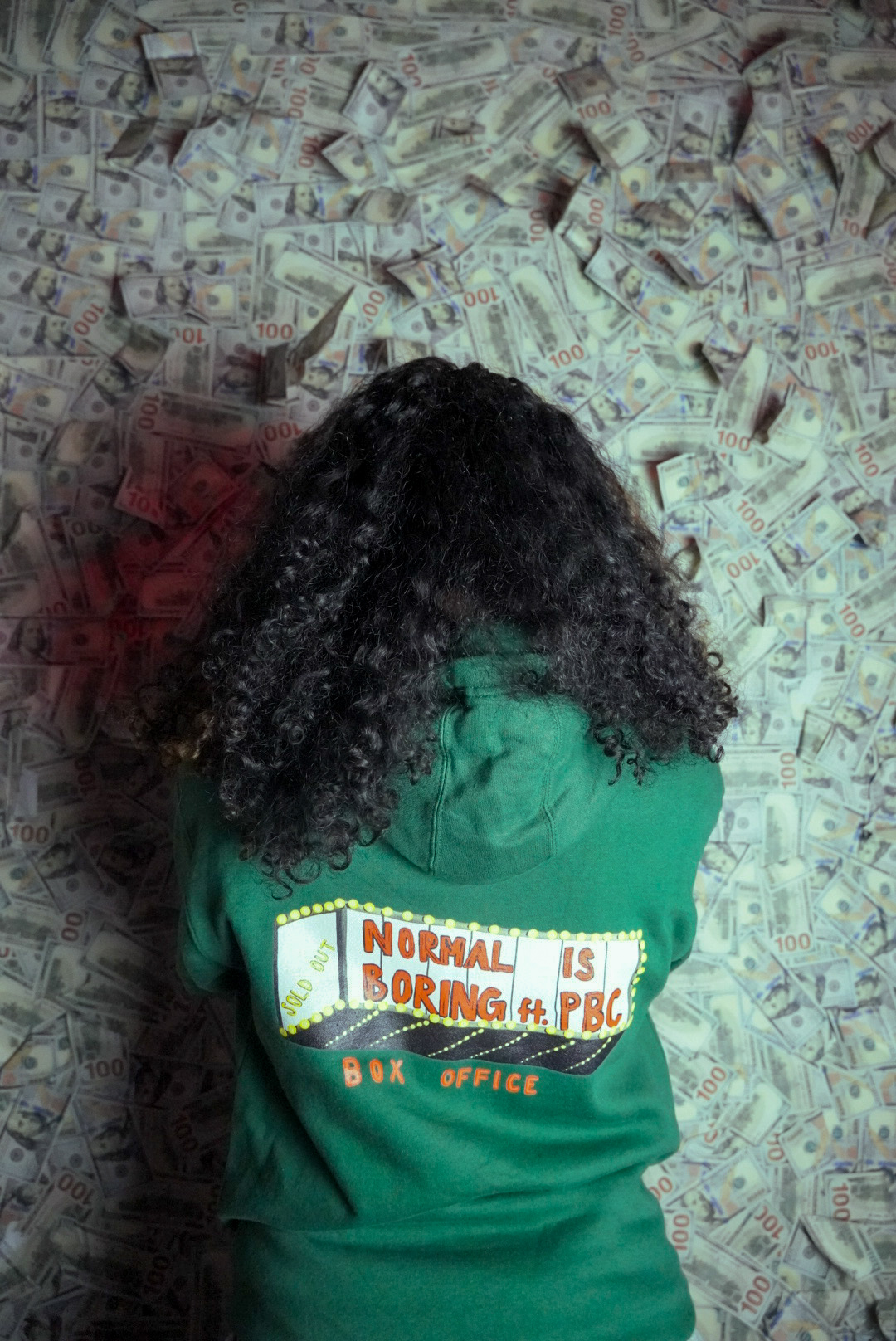 Person with curly hair lying on a surface covered with dollar bills, wearing a green shirt with a colorful sign.