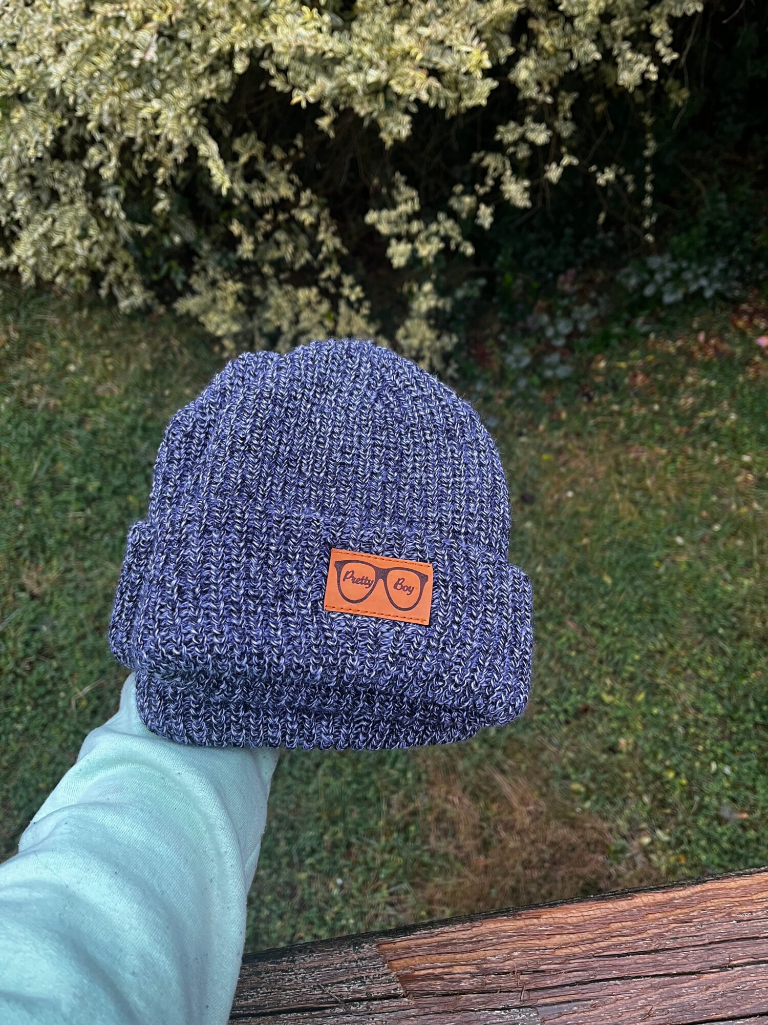 Person holding a blue knit beanie with a logo patch outdoors, grass and trees in background.