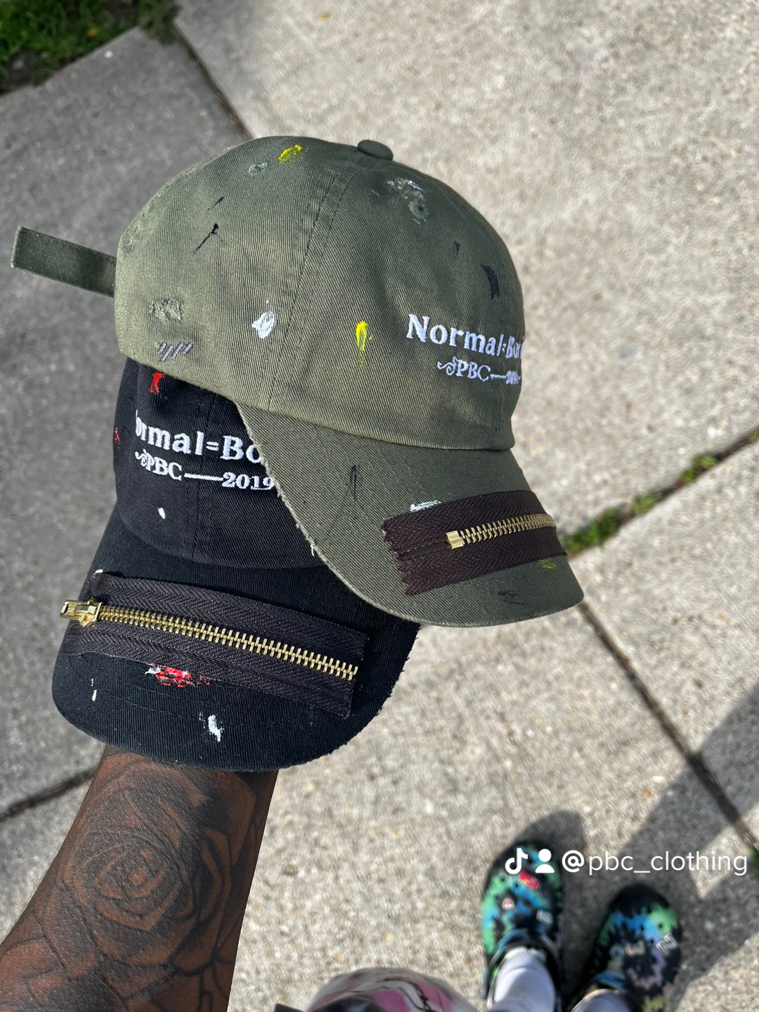 Green baseball cap with yellow accents and text, black bag with zipper, and part of a person's arm and shoes on sidewalk.
