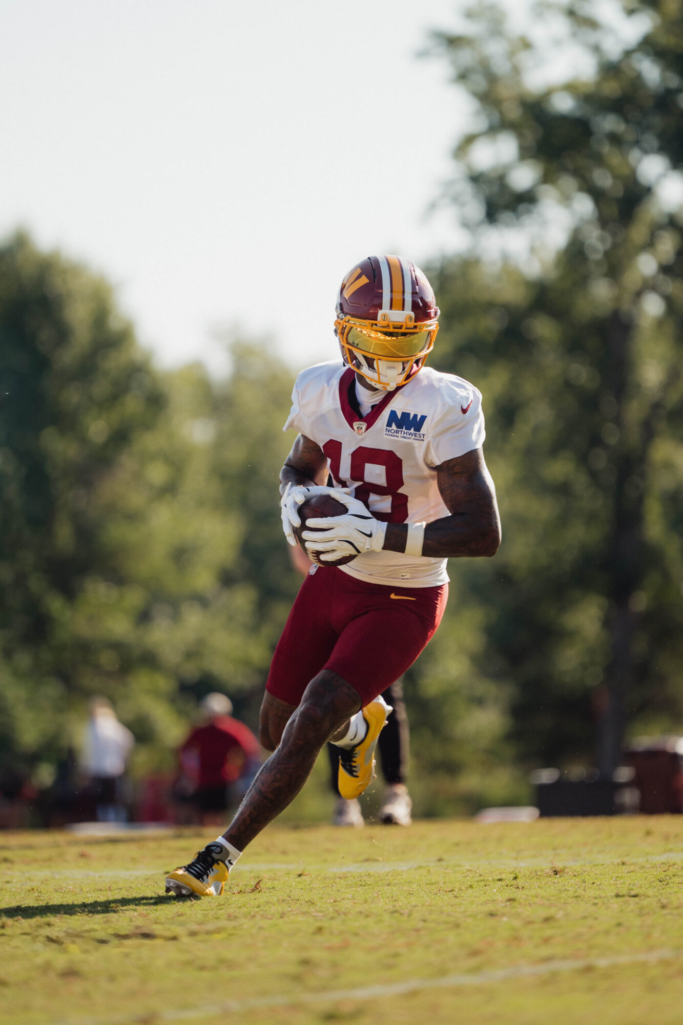 Football player running on field with trees in background, wearing maroon pants and white jersey with number 18.