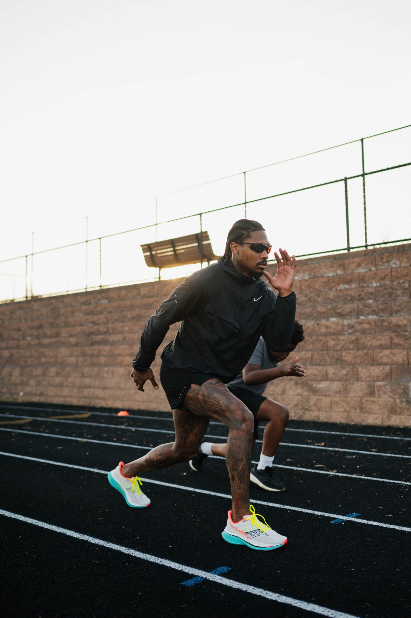 Person running on track wearing sunglasses, black jacket, colorful running shoes, with a brick wall and fence in background.