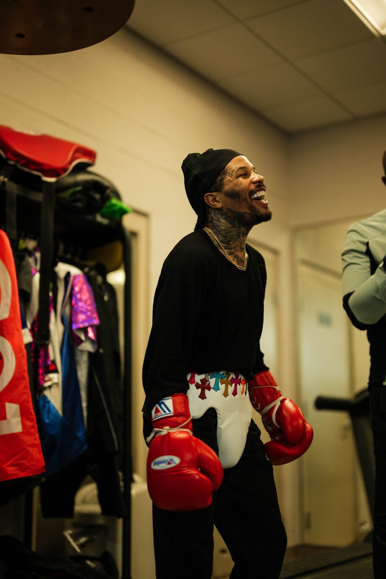 Person wearing boxing gloves and a black head covering, smiling in a room with clothing and equipment.