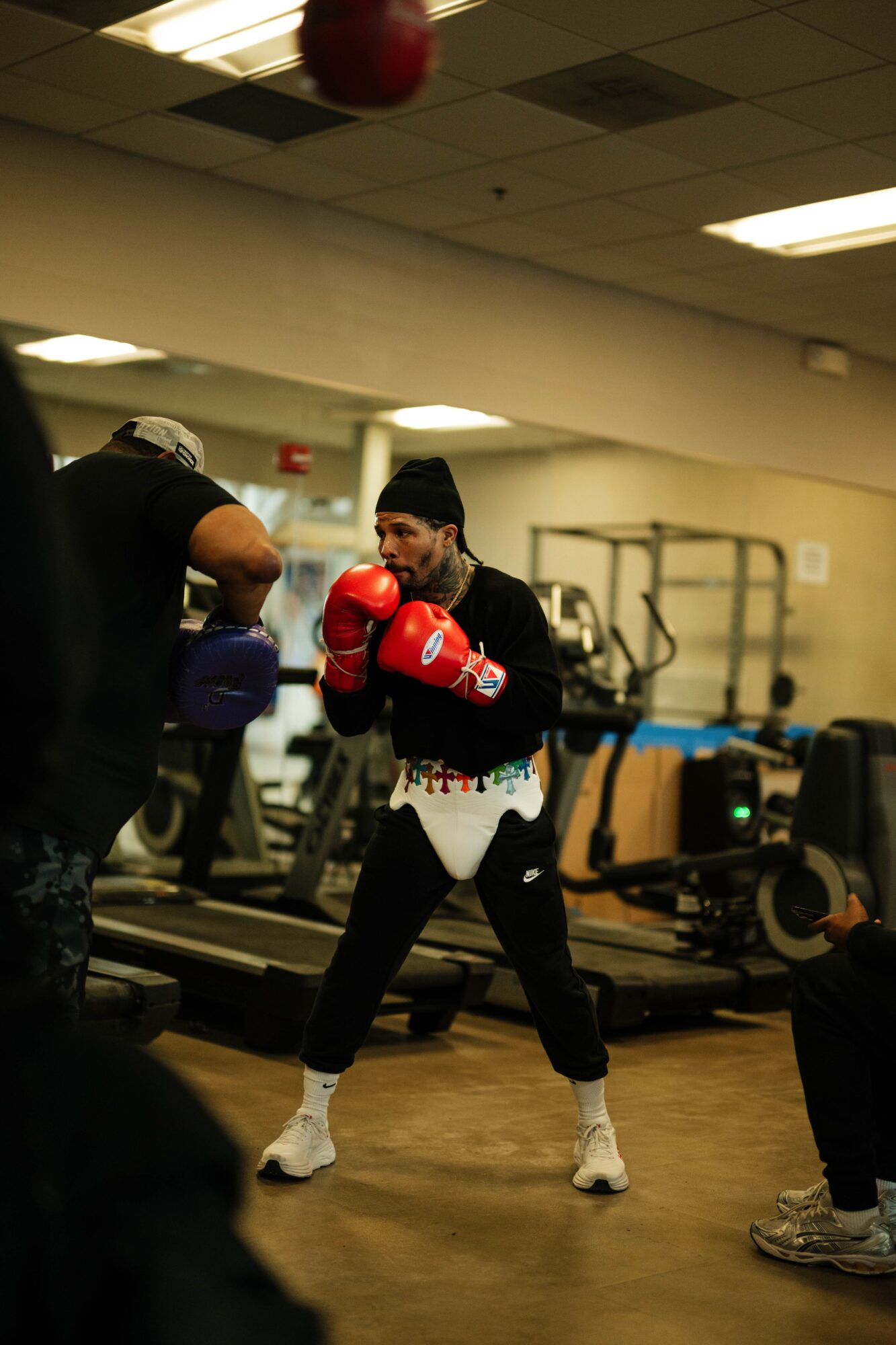 Woman in boxing gloves practicing in gym, surrounded by equipment and other people.