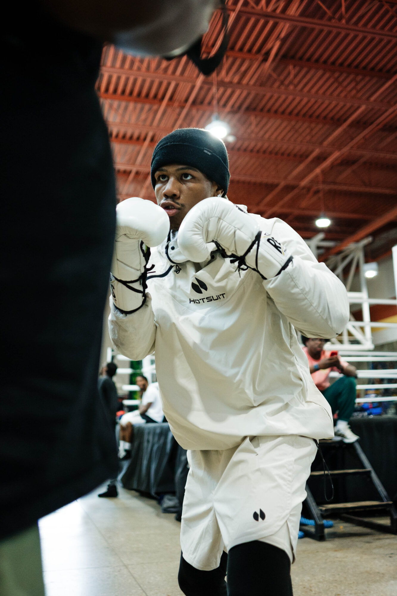 Young boxer in white gear and black beanie practicing boxing in a gym, with people and equipment in background.