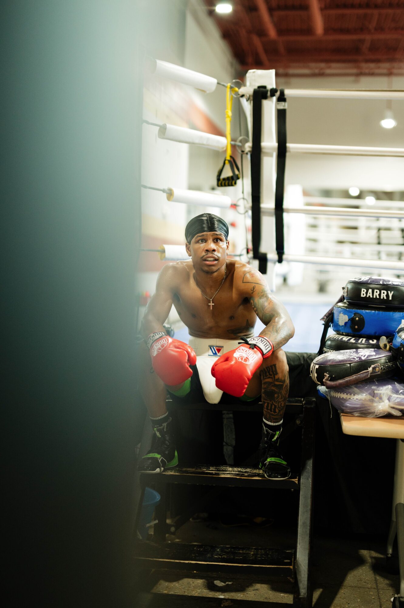 Boxer sitting on a stool in a gym, wearing red boxing gloves and black shorts, with boxing equipment nearby.