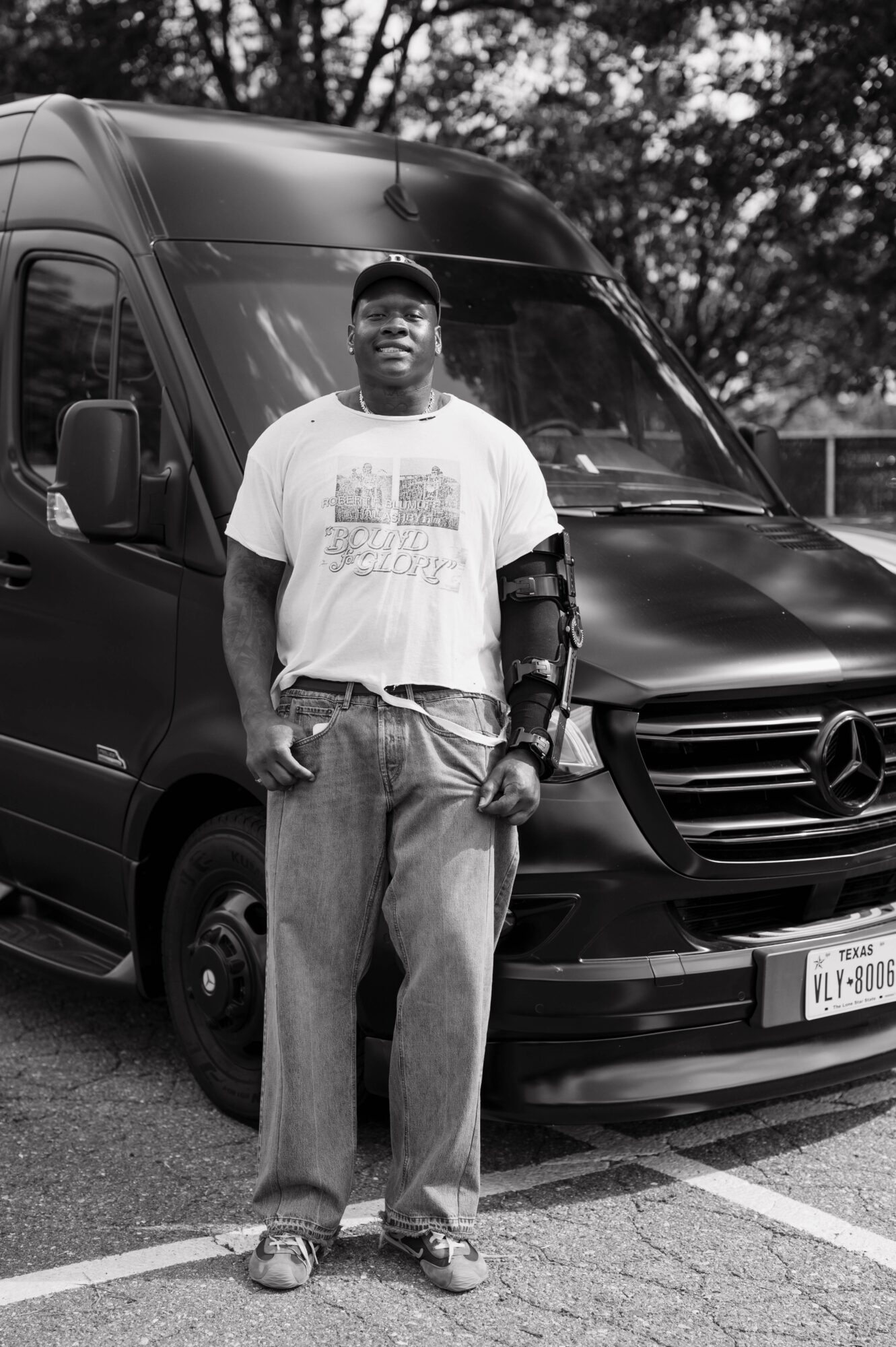 Man standing in front of a van, wearing a cap, t-shirt, and jeans, with trees in the background.