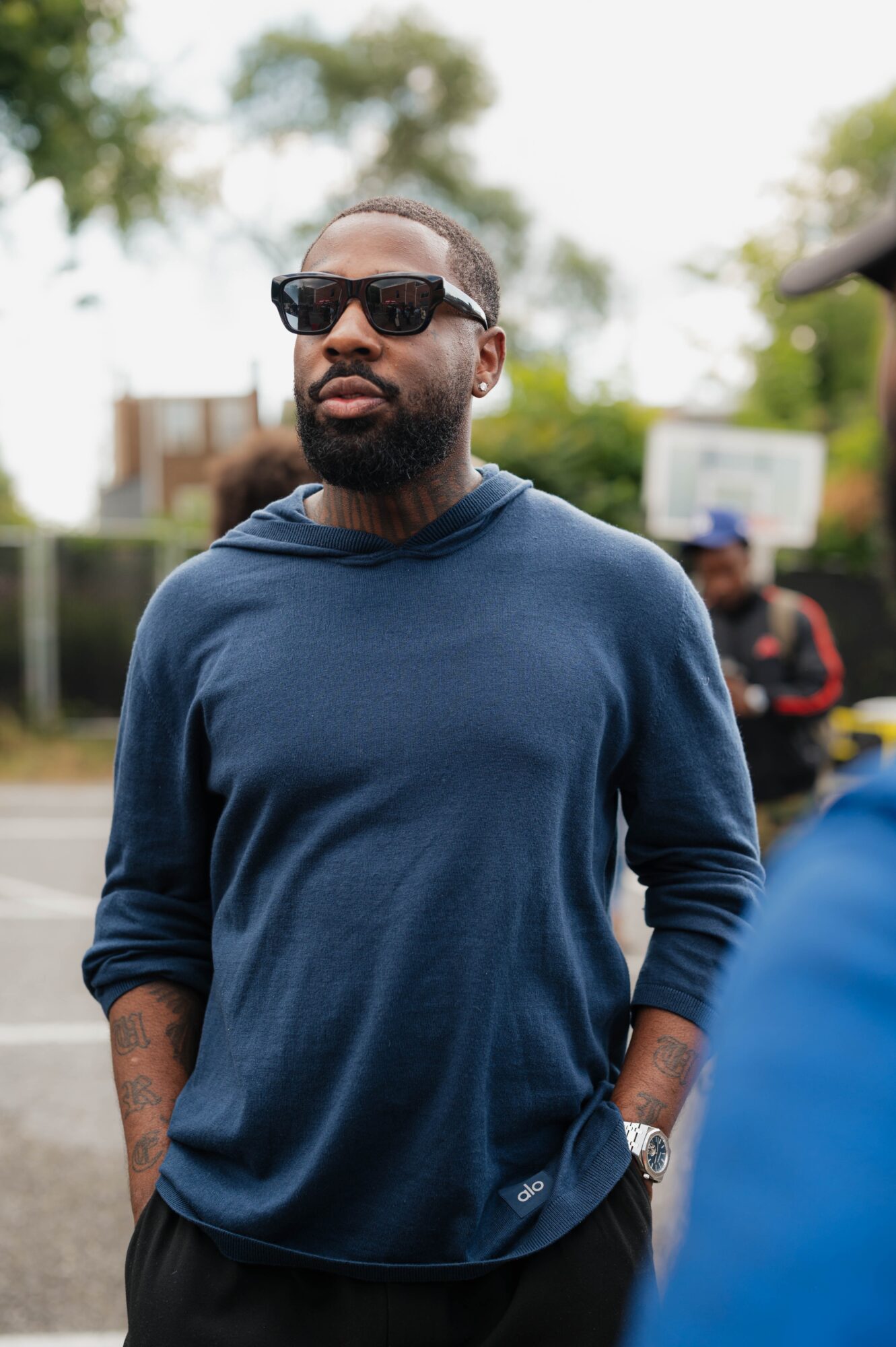 Man with sunglasses and beard wearing a blue hoodie outdoors, trees and a basketball hoop in background.